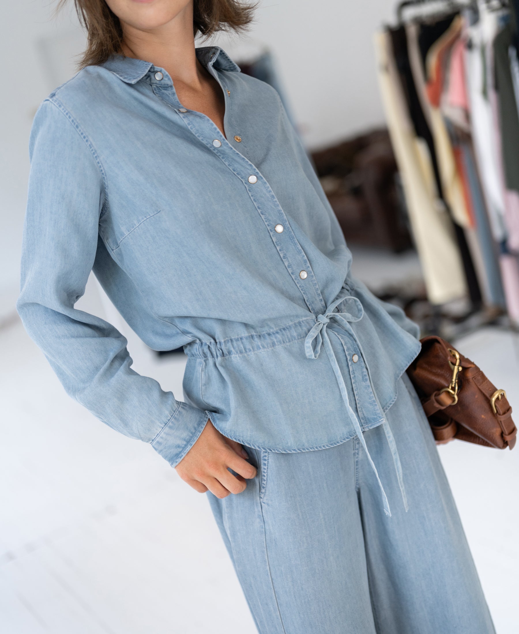 Woman wearing bleached Tencel denim blouse, standing with clothes displayed in the background.
