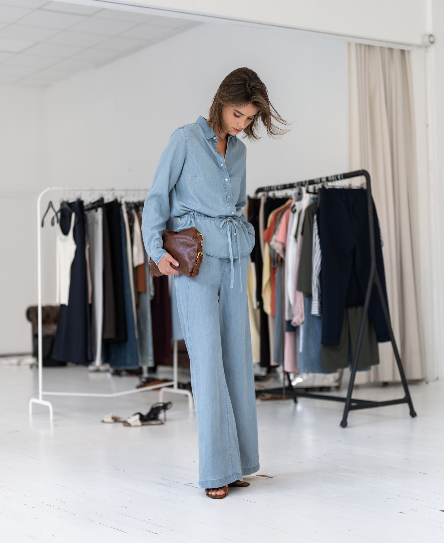 Woman wearing a bleached Tencel denim blouse stands in a studio with clothing racks in the background.