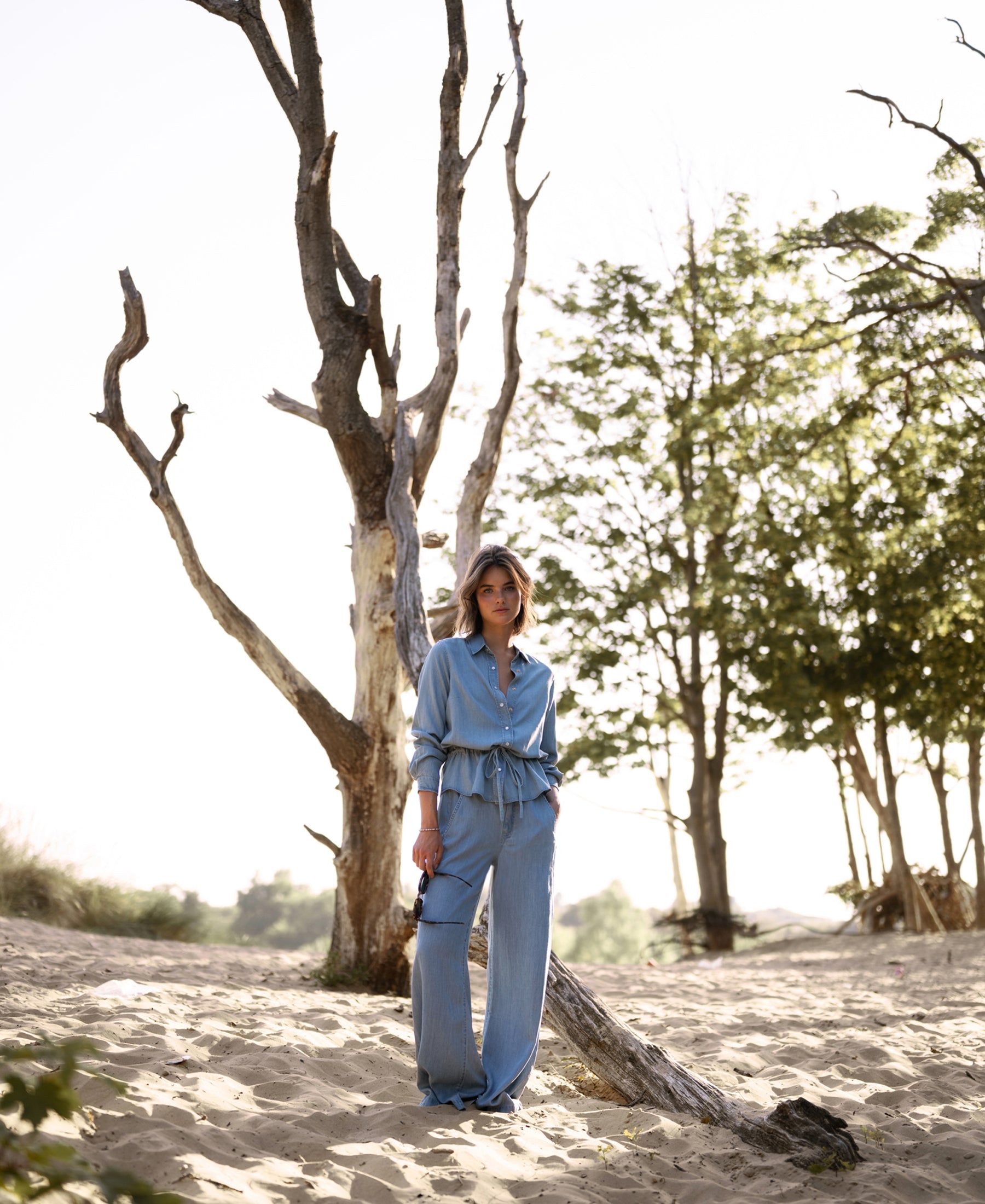 Woman wears light blue Tencel denim jeans on sandy ground near a tall, leafless tree.