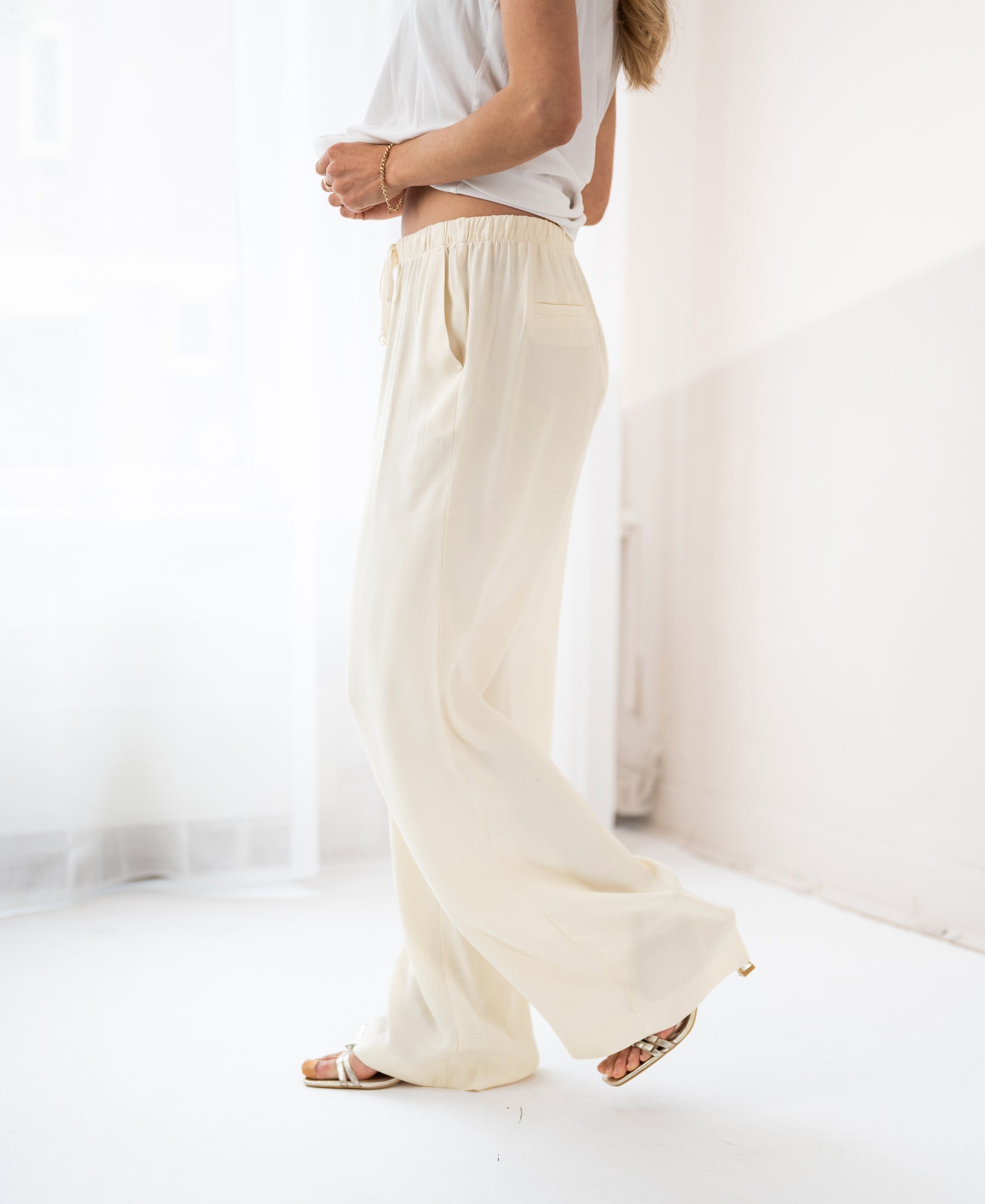 Cream-colored LA LYNETTE pants displayed in a bright, minimalistic white room.