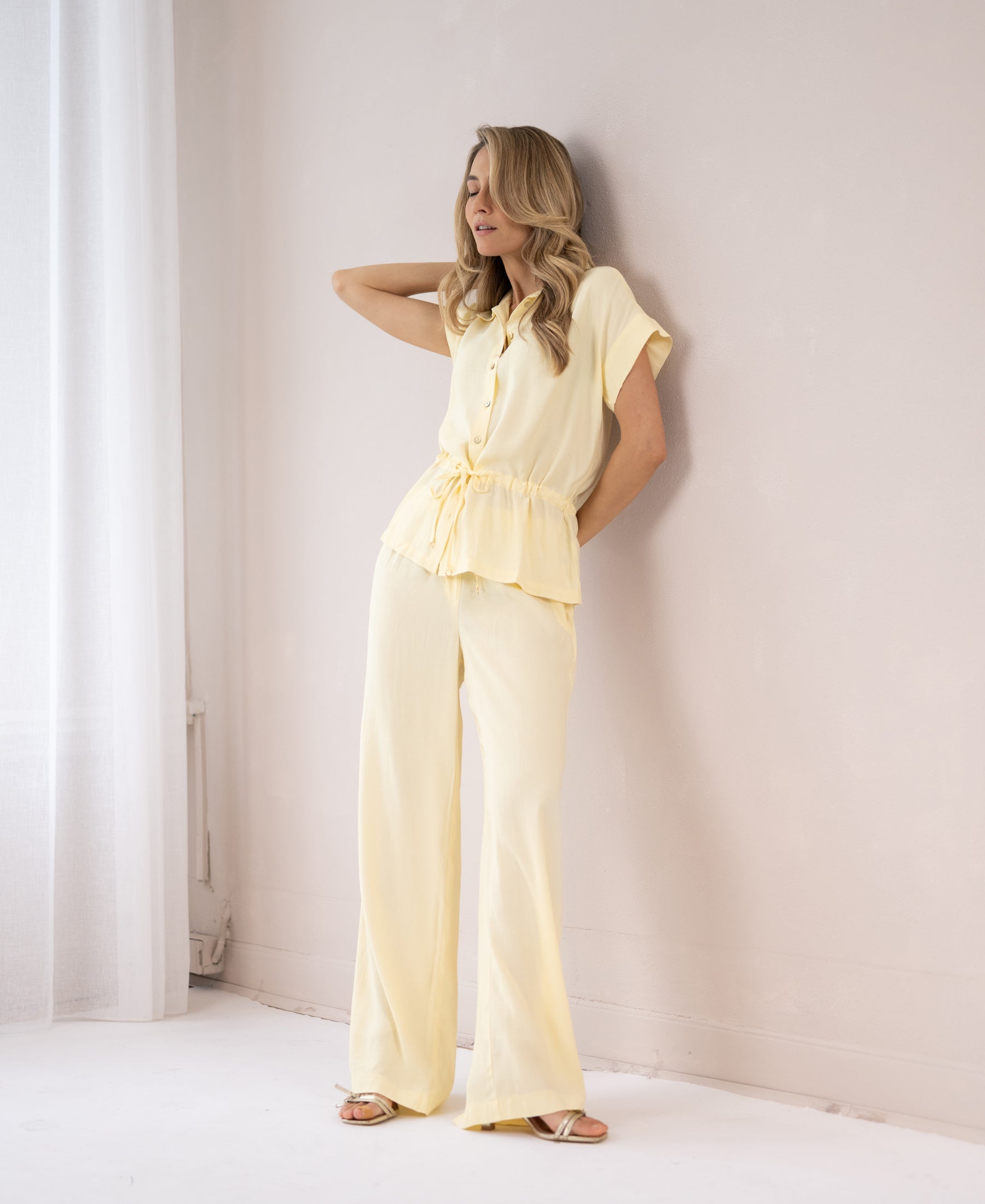 Light yellow blouse in soft fabric, shown on a woman standing by a white wall with eyes closed, in gentle lighting.