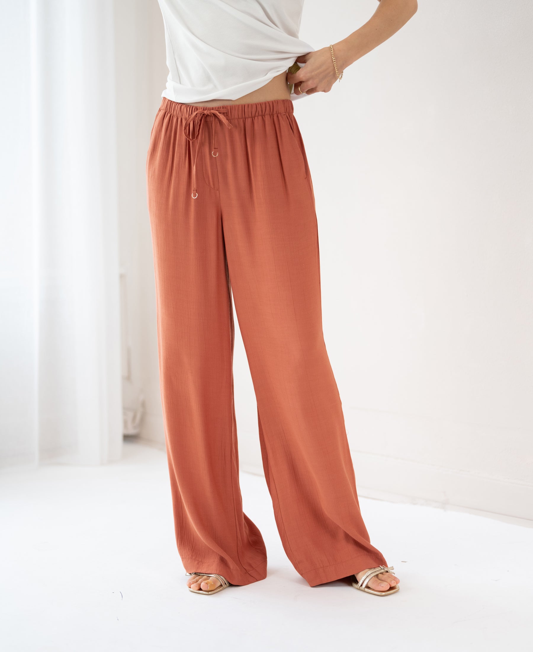 Terra-colored LA LYNETTE pants by PLEIN PUBLIQUE shown in a minimal, bright room with white walls.