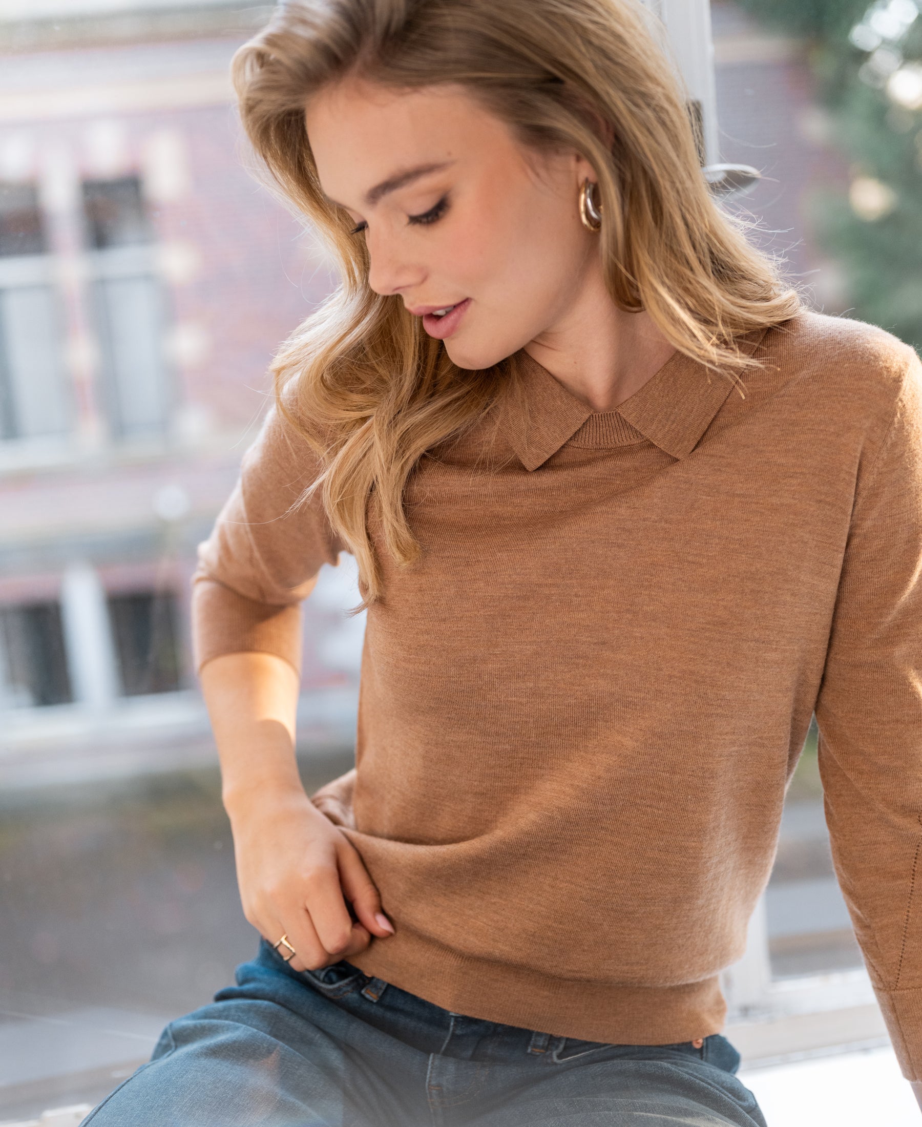 Woman wearing a merino wool polo sweater in Chai Tea color sits by a window, smiling gently.