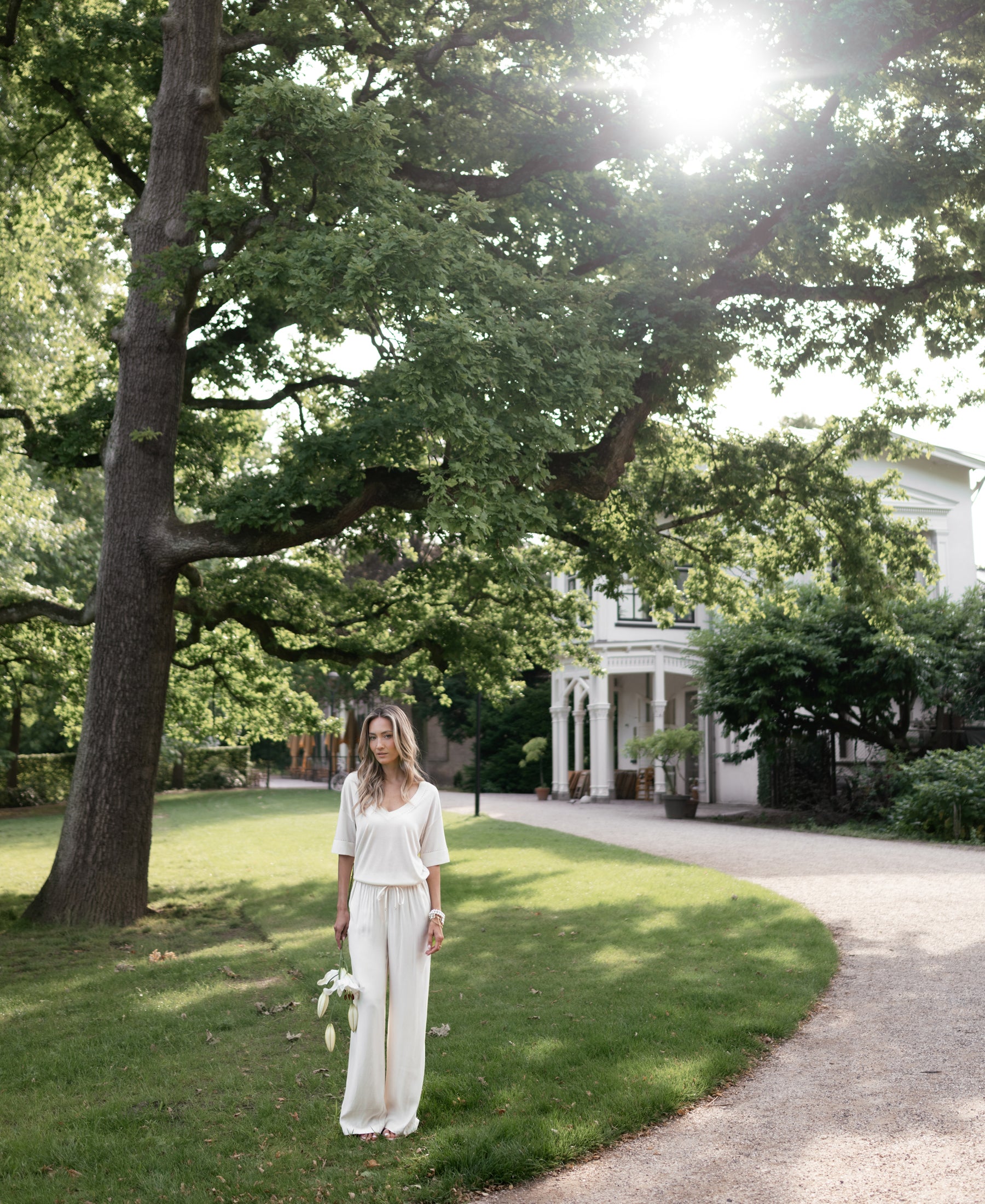 Woman wearing cream Plein Publique LA Lynette pants stands on a lawn in sunlight near a tree and white house.