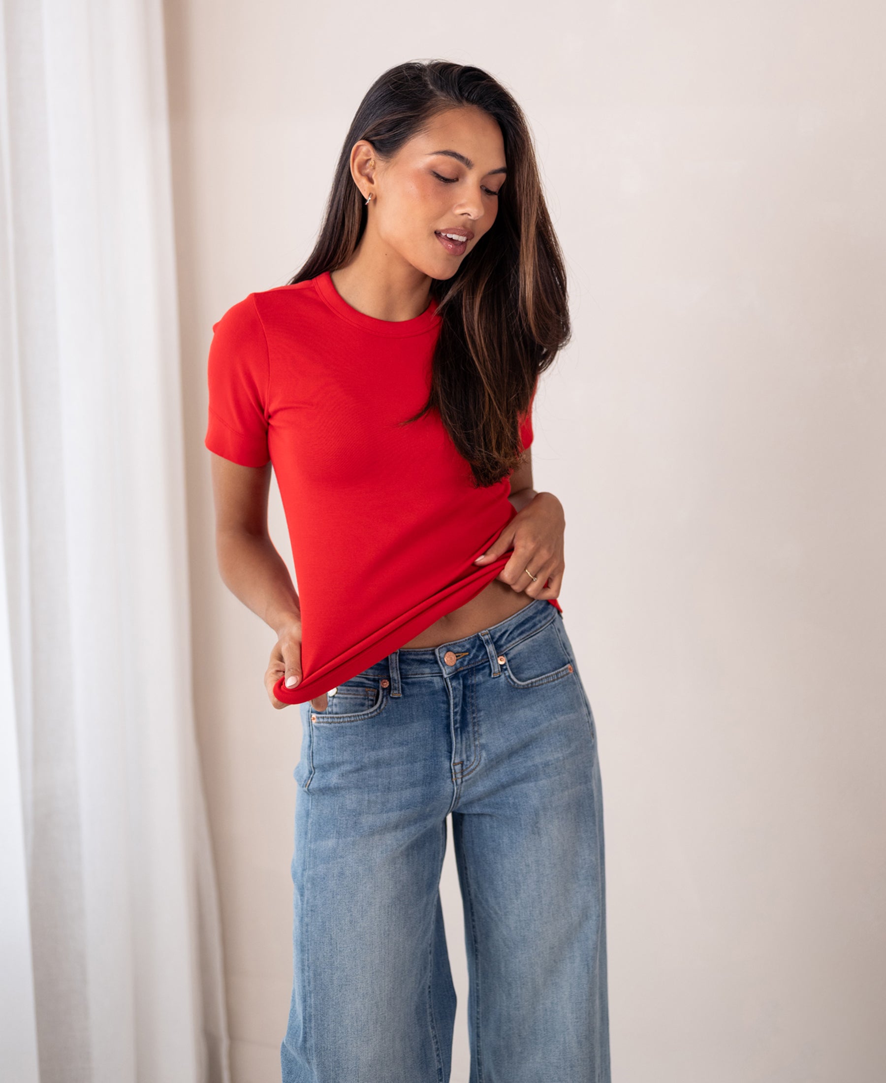 Woman wearing a Plein Publique Luxe & Soft red T-shirt indoors, looking down and holding the hem of the shirt.