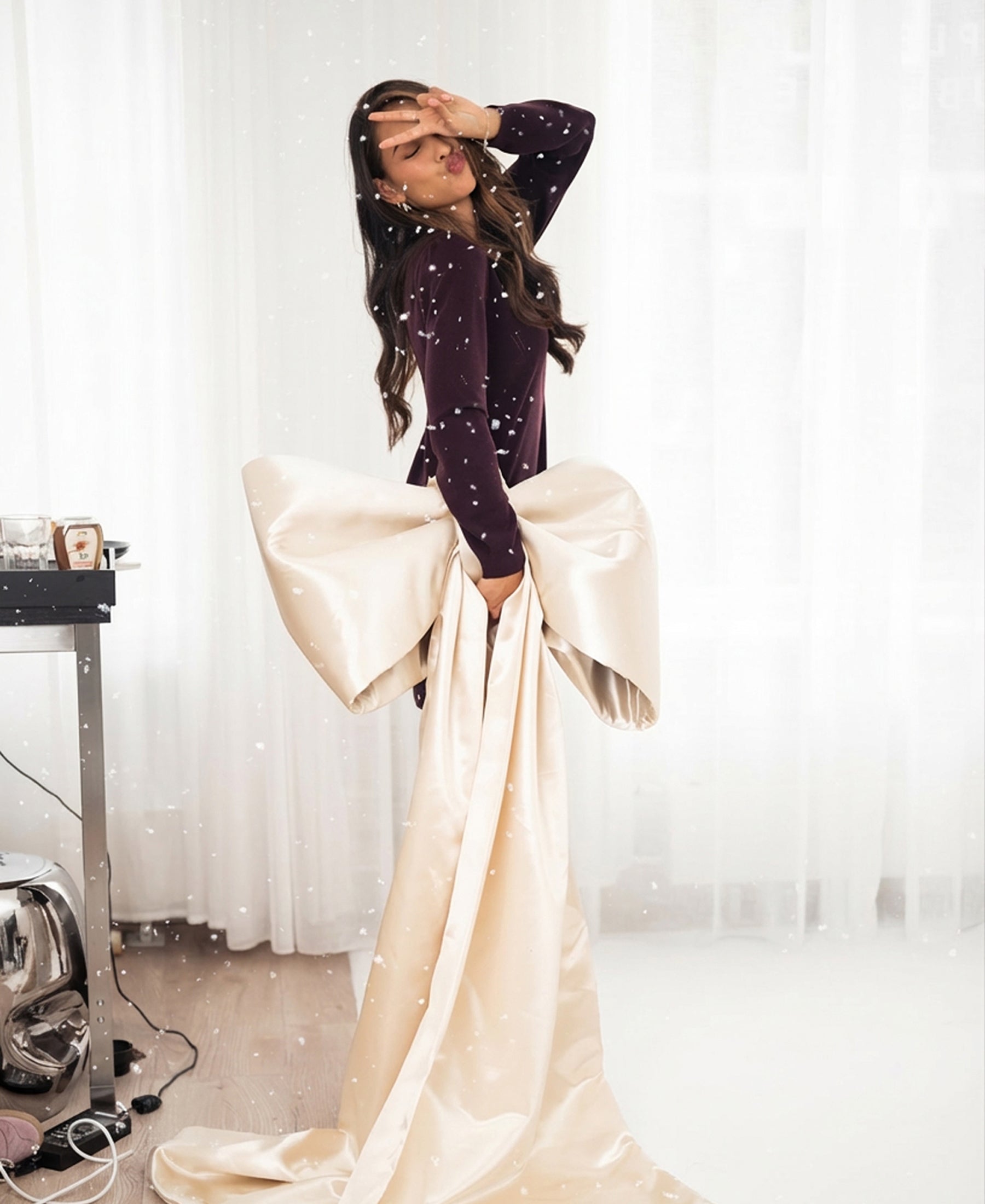 Woman poses indoors in an aubergine merino wool dress, hand raised, with white curtains in the background.