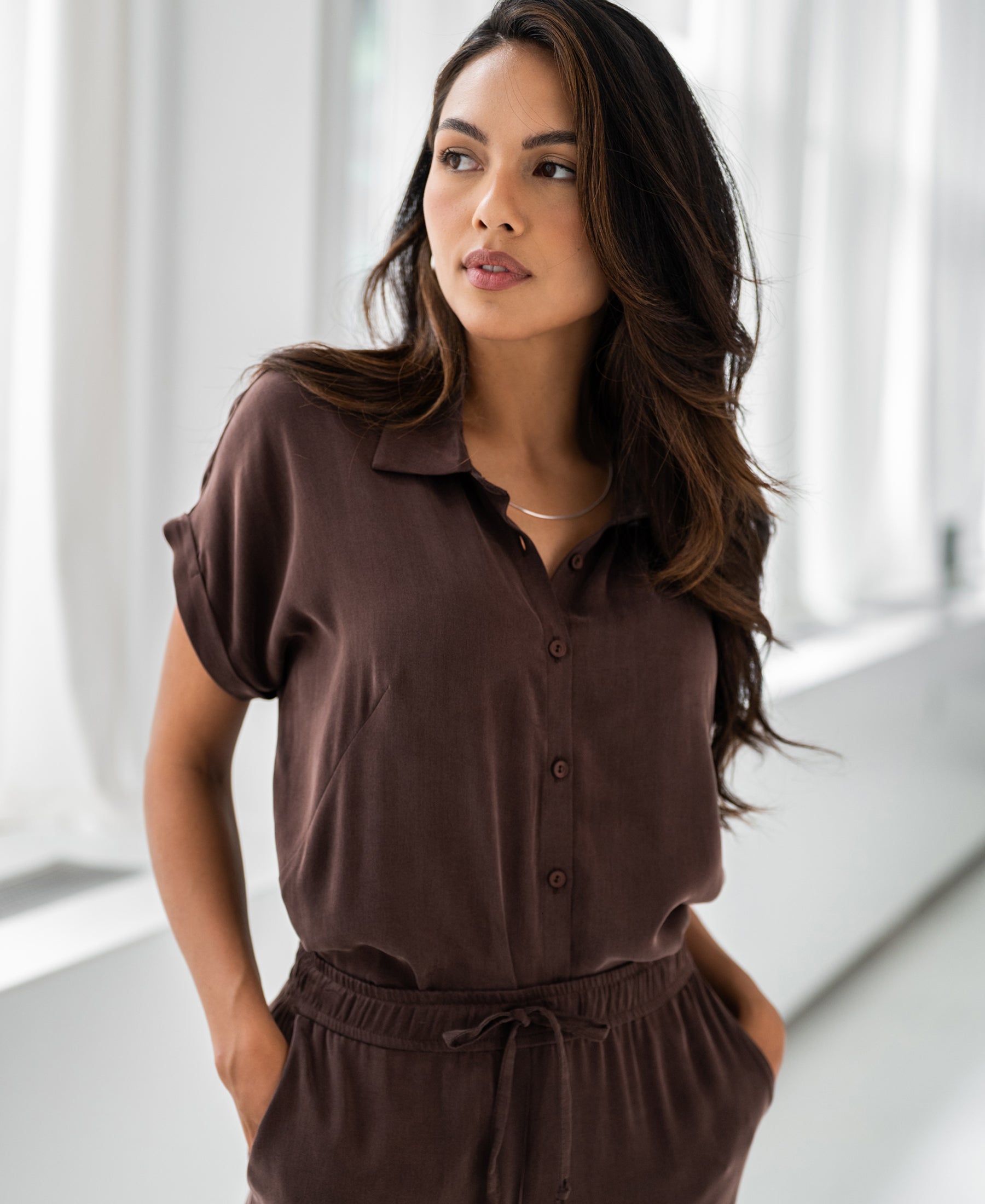 Woman wearing the Plein Publique cupro blouse in brown stands indoors by bright windows, looking to the side.
