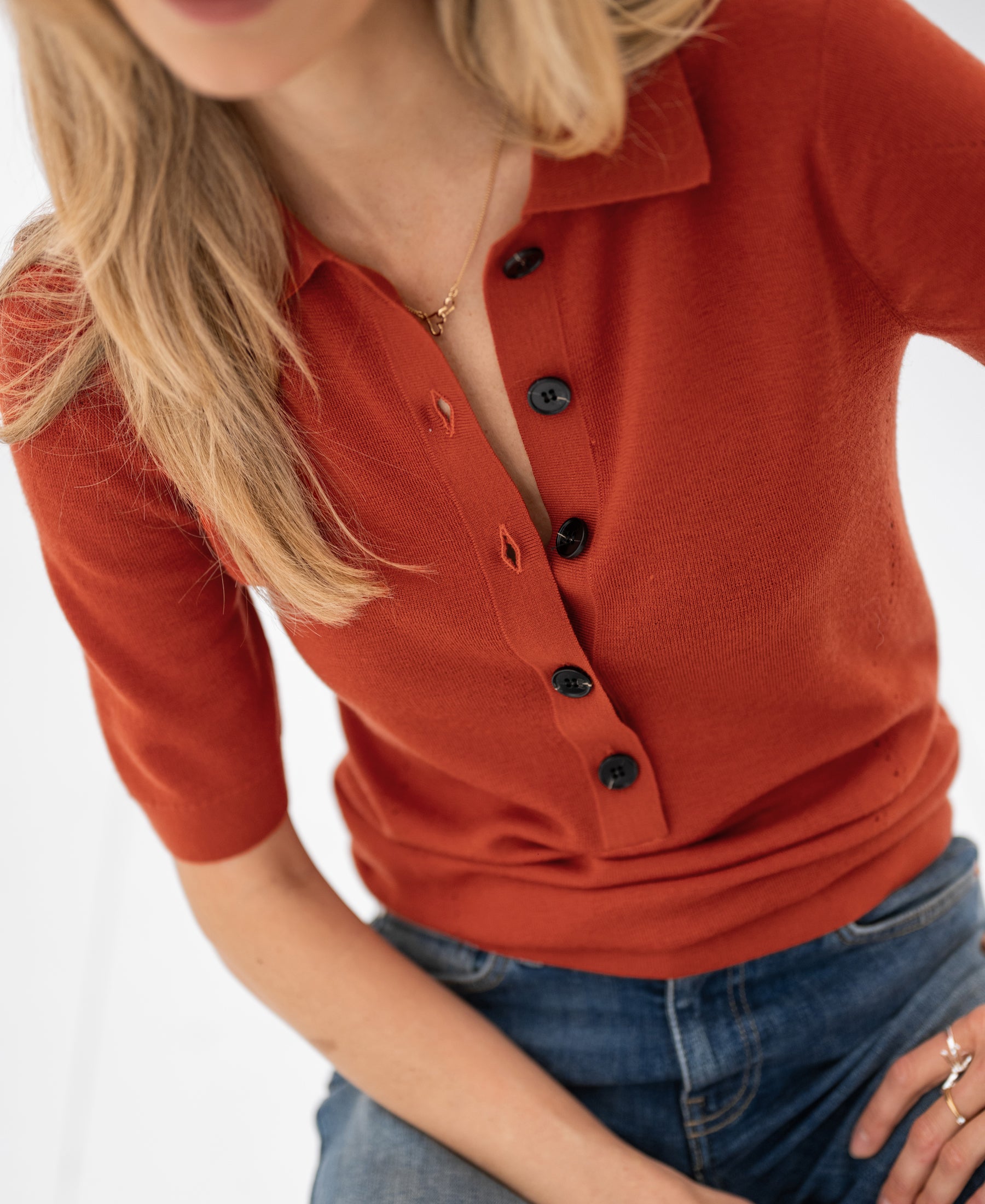 Woman wears the PLEIN PUBLIQUE merino wool polo shirt in cayenne, seated with one arm resting on her knee.
