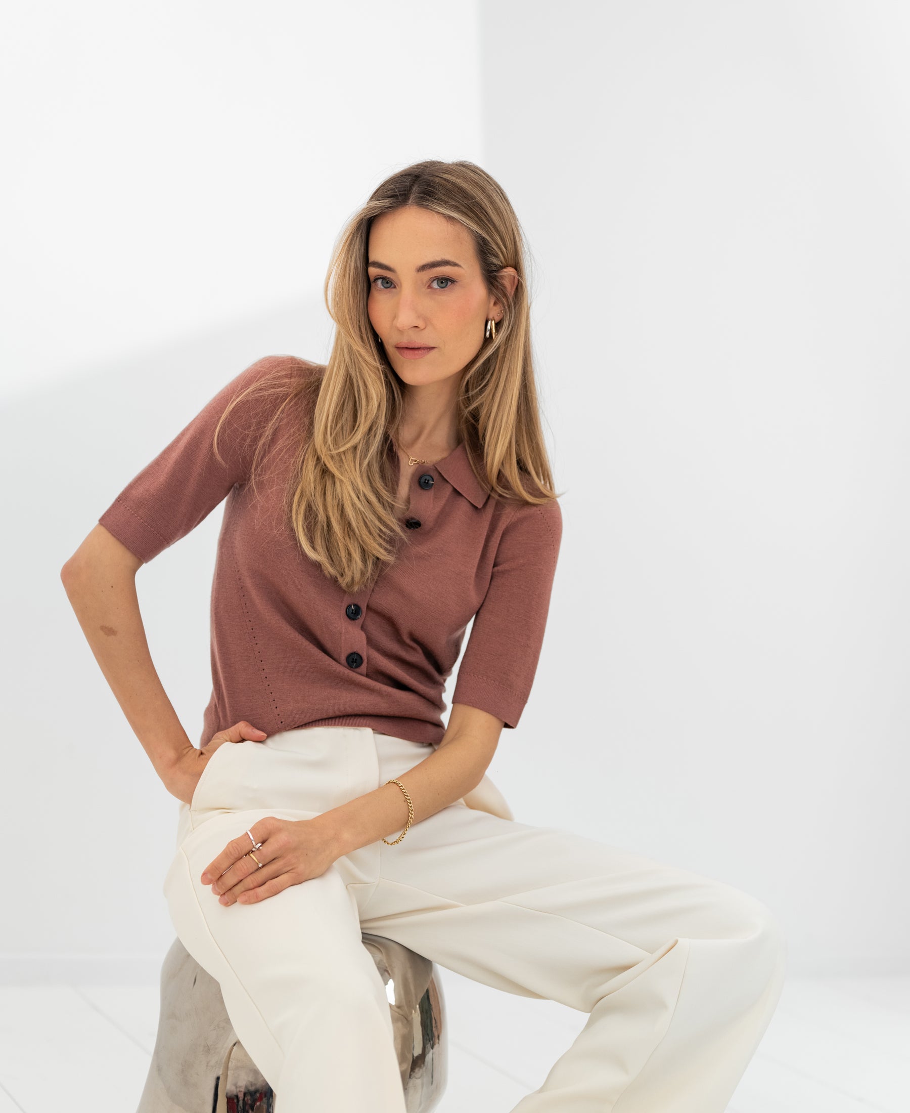 Woman wearing a dusty pink and white merino wool polo sits on a stool.