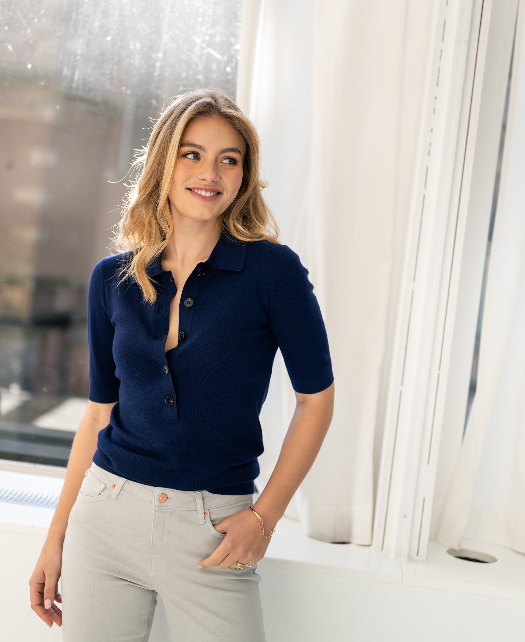 Navy merino wool polo sweater, all-season classic, shown on model standing by a window.