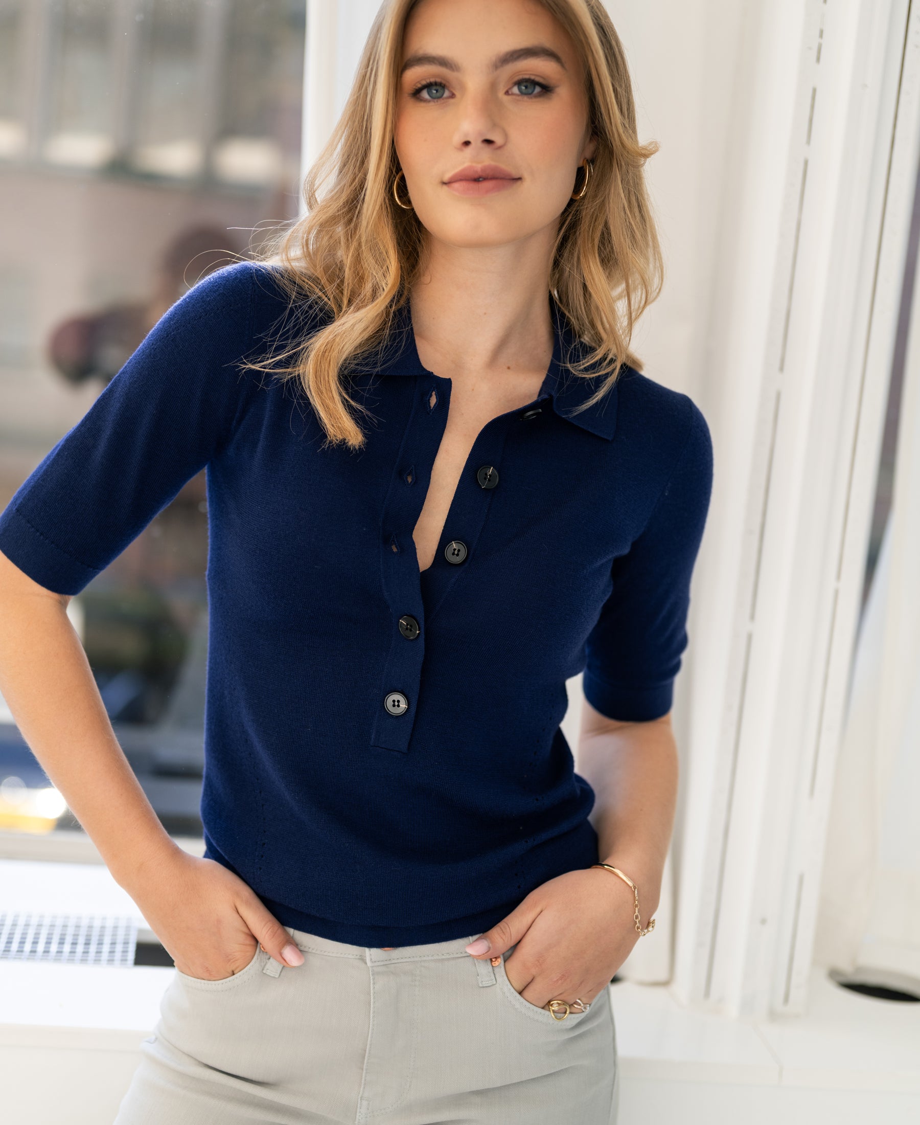 Navy merino wool polo shirt, front view, shown by a woman standing by a window with hands in pockets, looking at the camera.