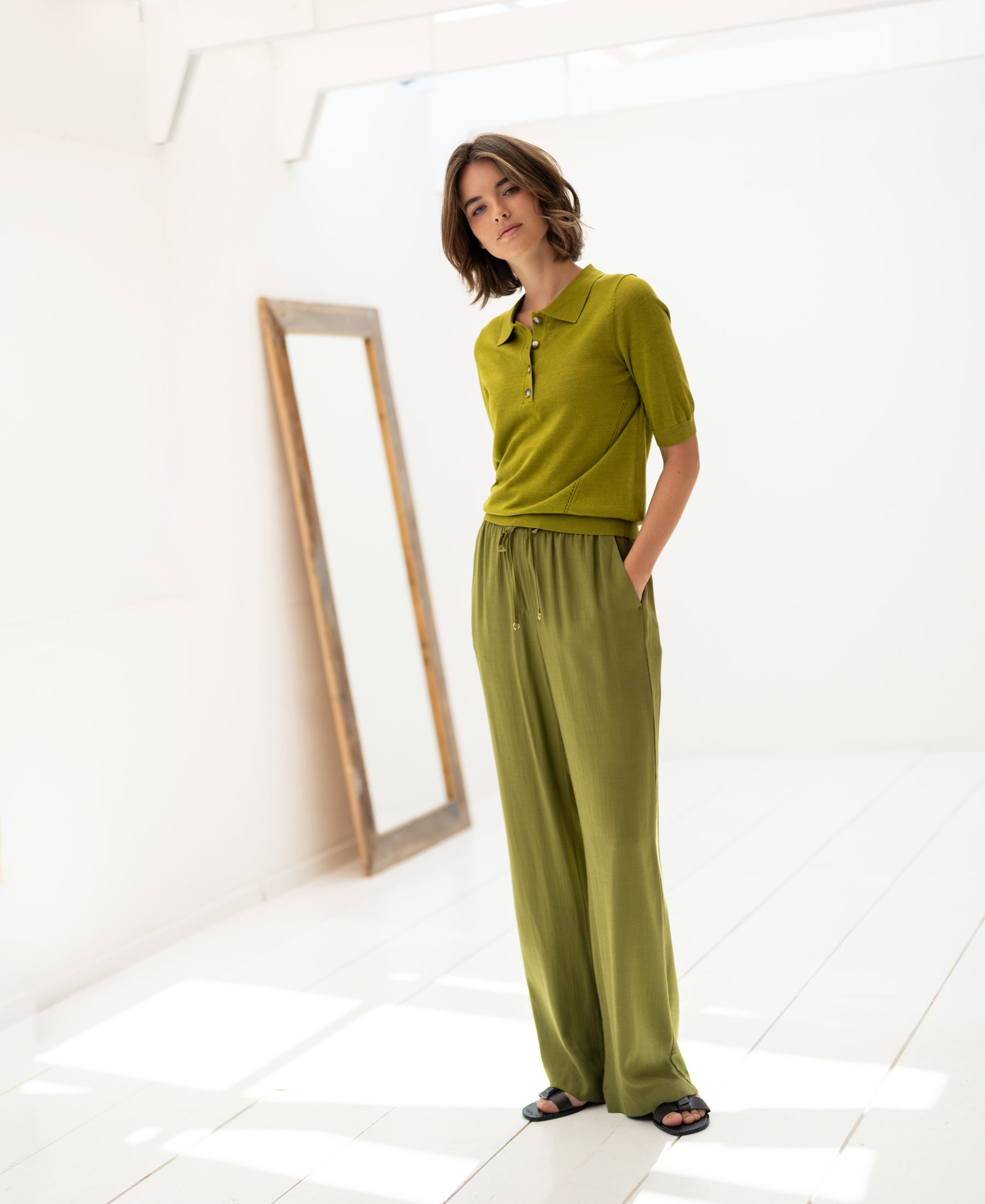 Olive green Merino wool-silk polo shirt shown on a woman in a bright, minimalist room with a floor mirror.