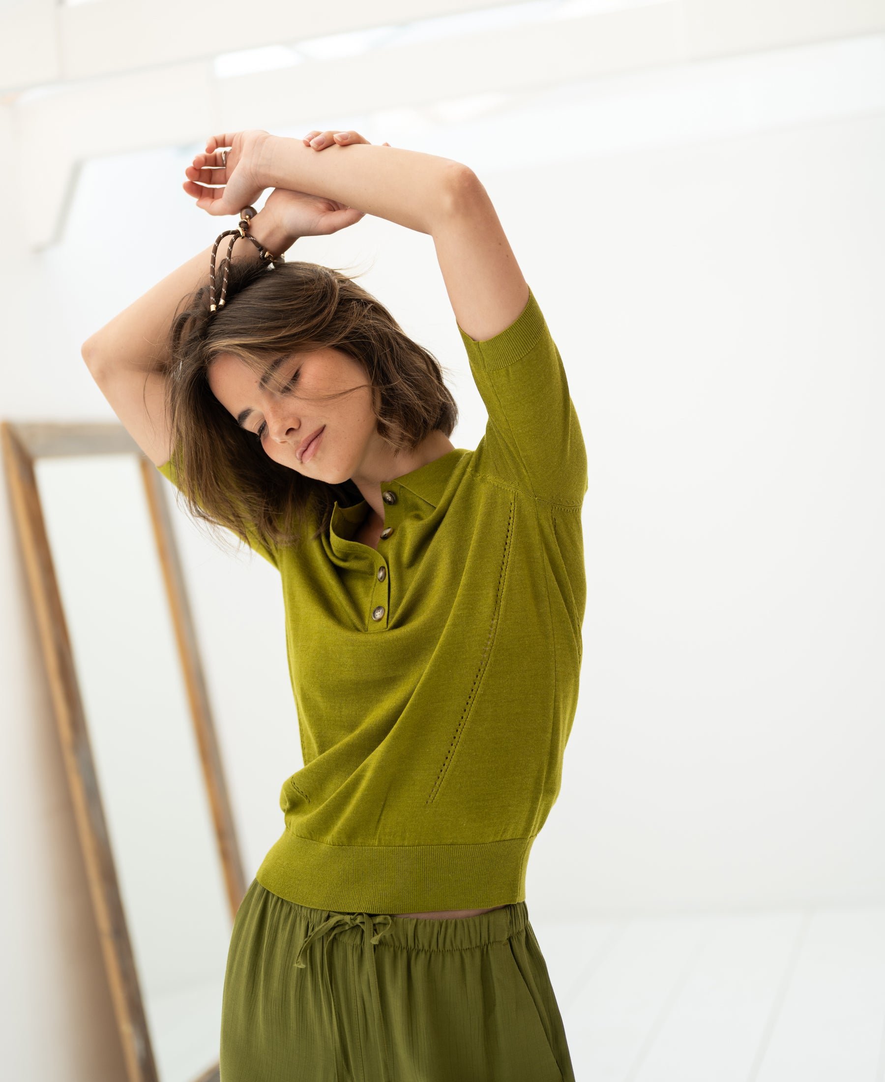 Woman wearing an olive green Merino-silk polo shirt stands with arms raised, eyes closed, next to a mirror.