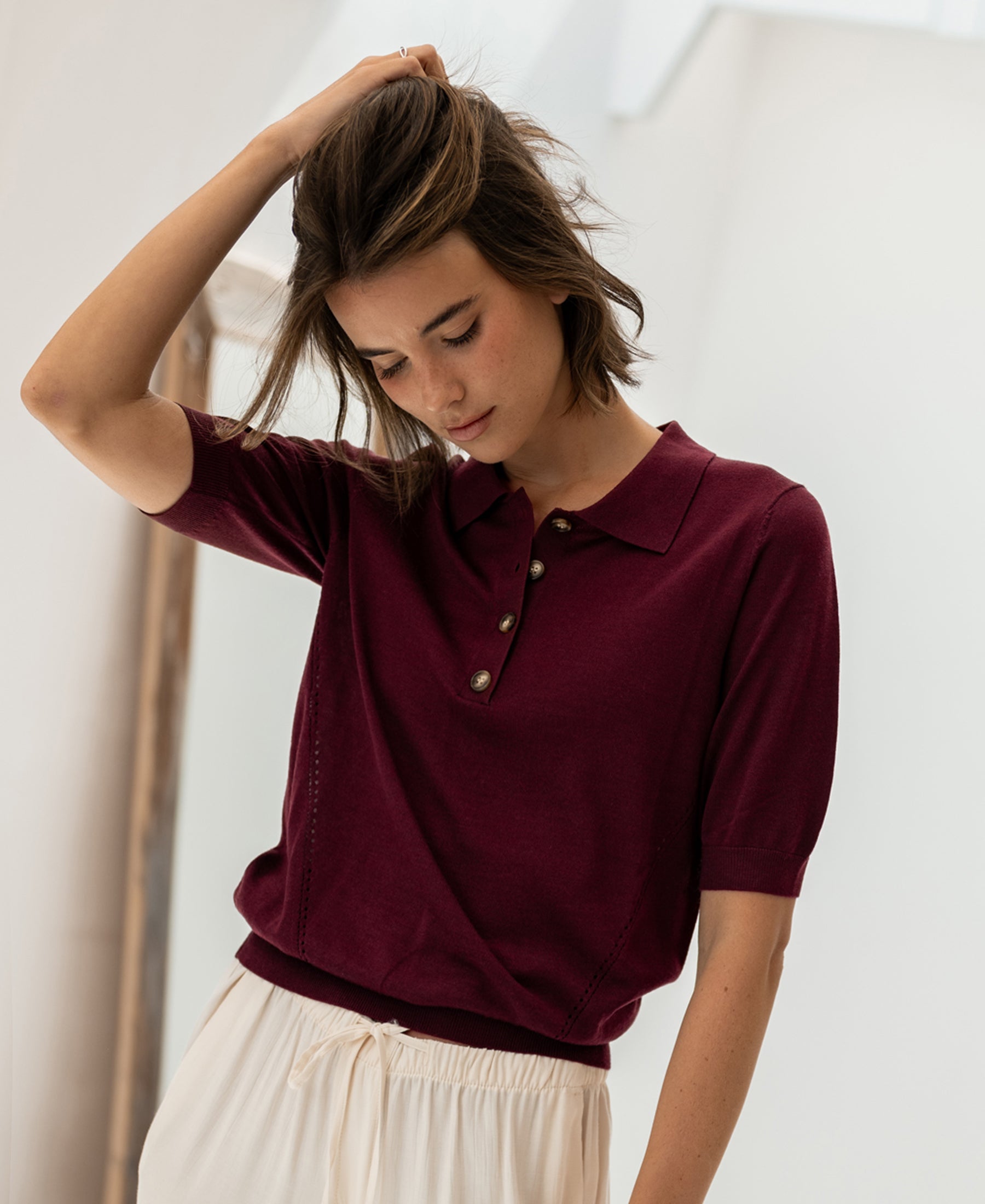 Woman wearing a plum Merino-silk LE PAULY polo by PLEIN PUBLIQUE stands indoors, looking down.