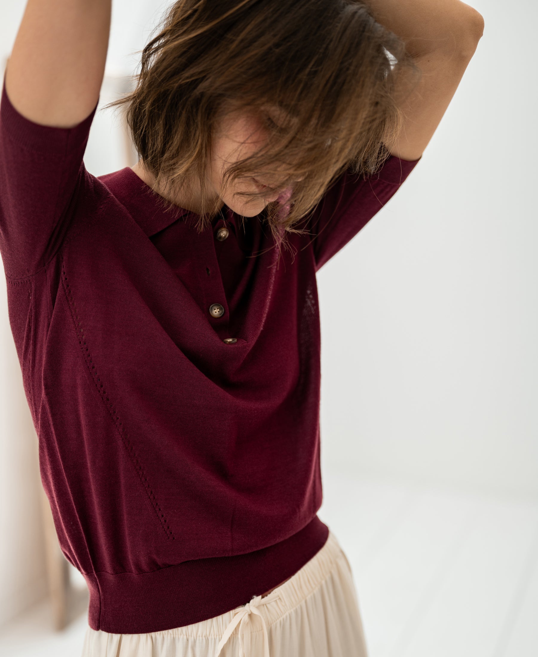 Plum and cream Merino wool-silk polo shirt, shown front view with the model standing, hands in hair, head down.