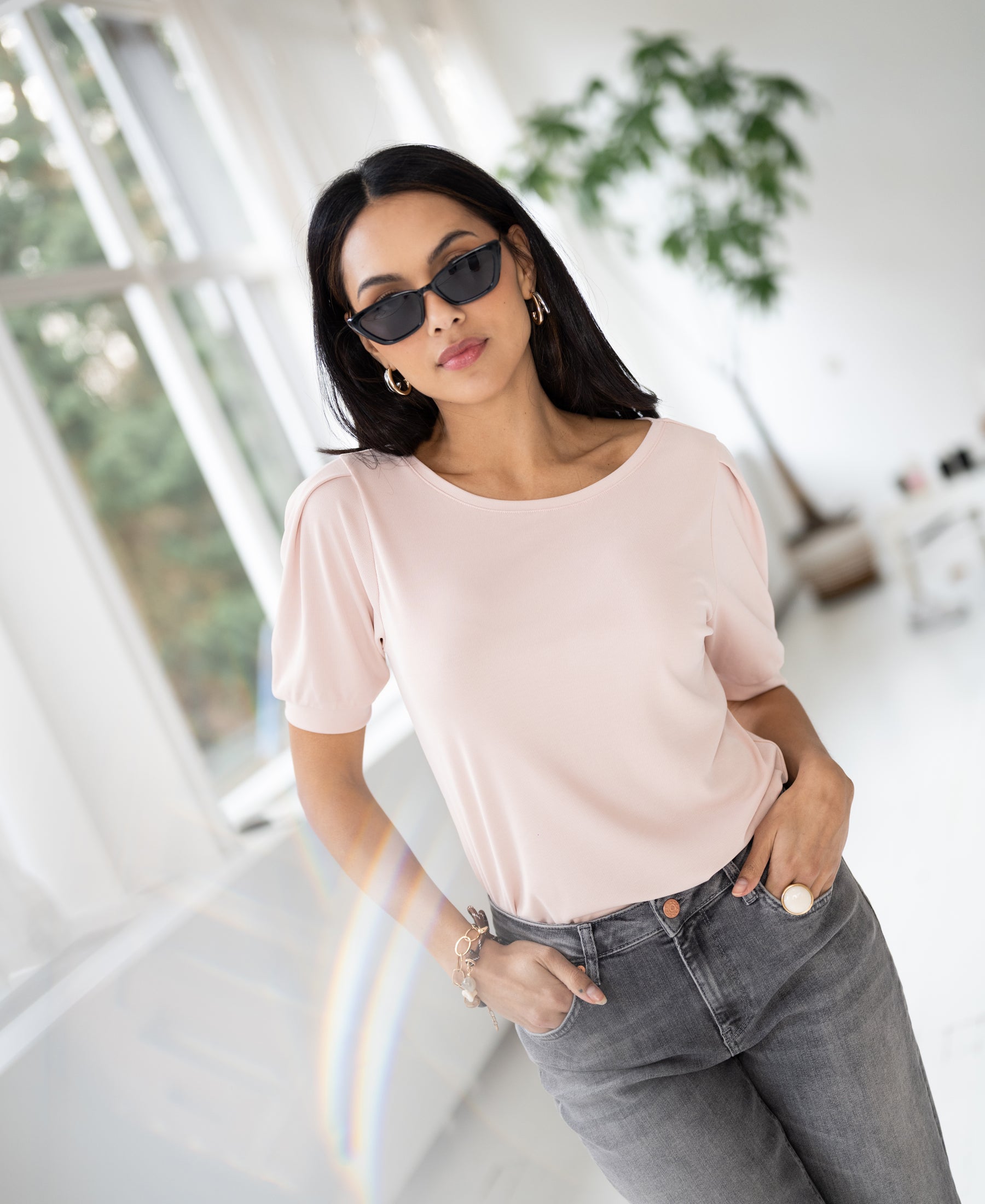 Woman wearing a blush ribbed T-shirt stands in a bright room with plants.