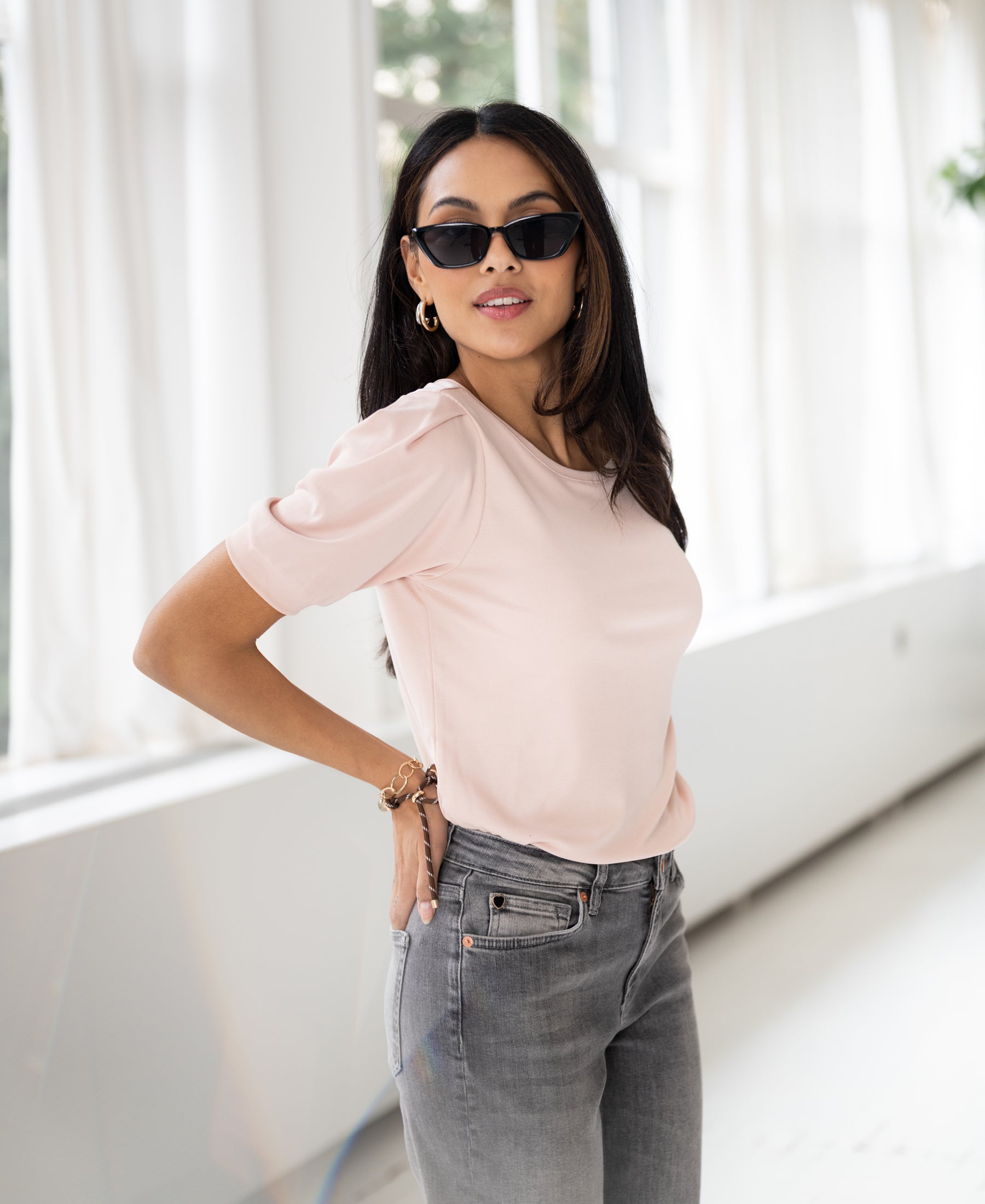 Woman wearing ribbed blush T-shirt stands by large bright windows.