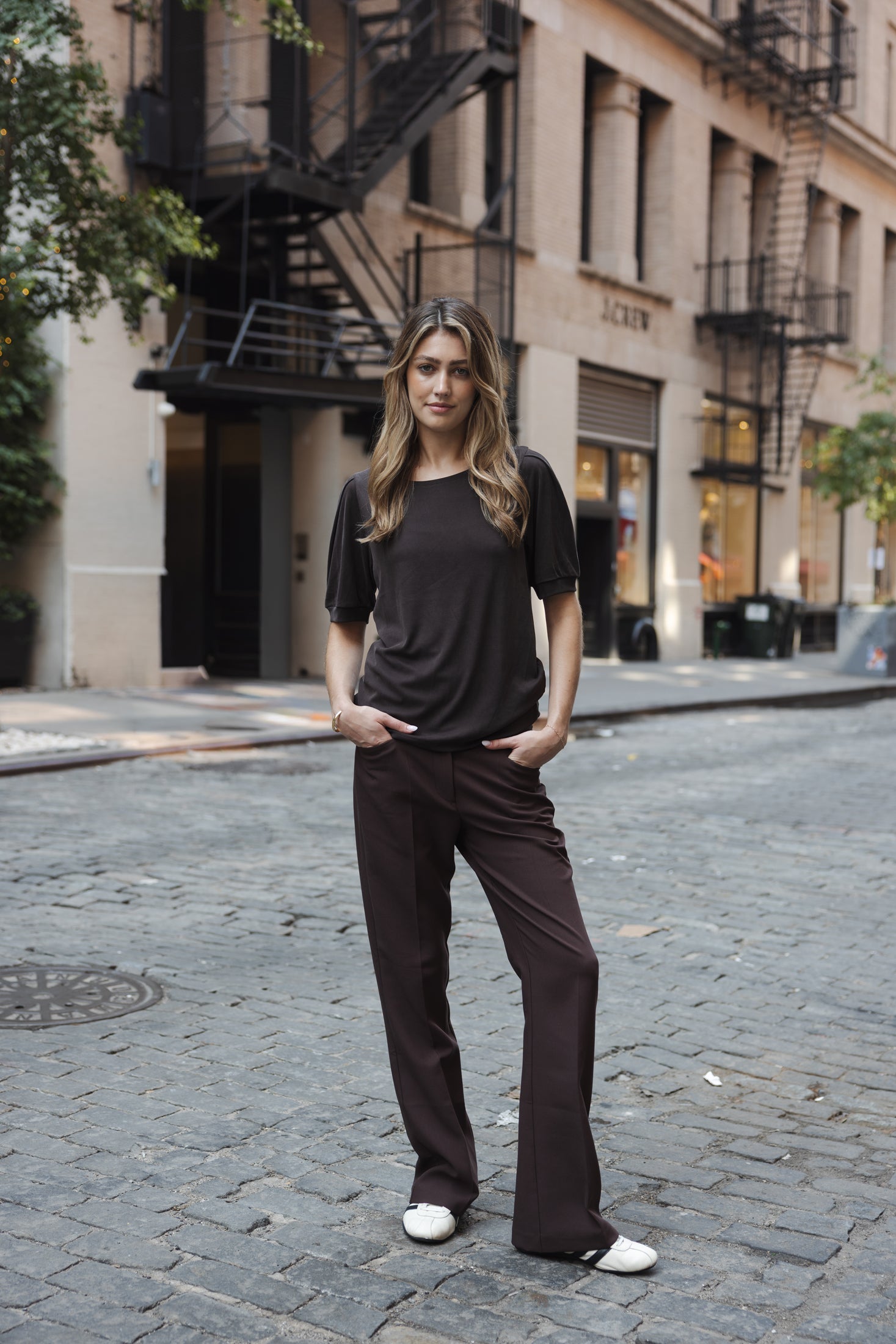 Woman wearing chocolate brown ribbed T-shirt, hands in pockets, standing on cobblestone street.