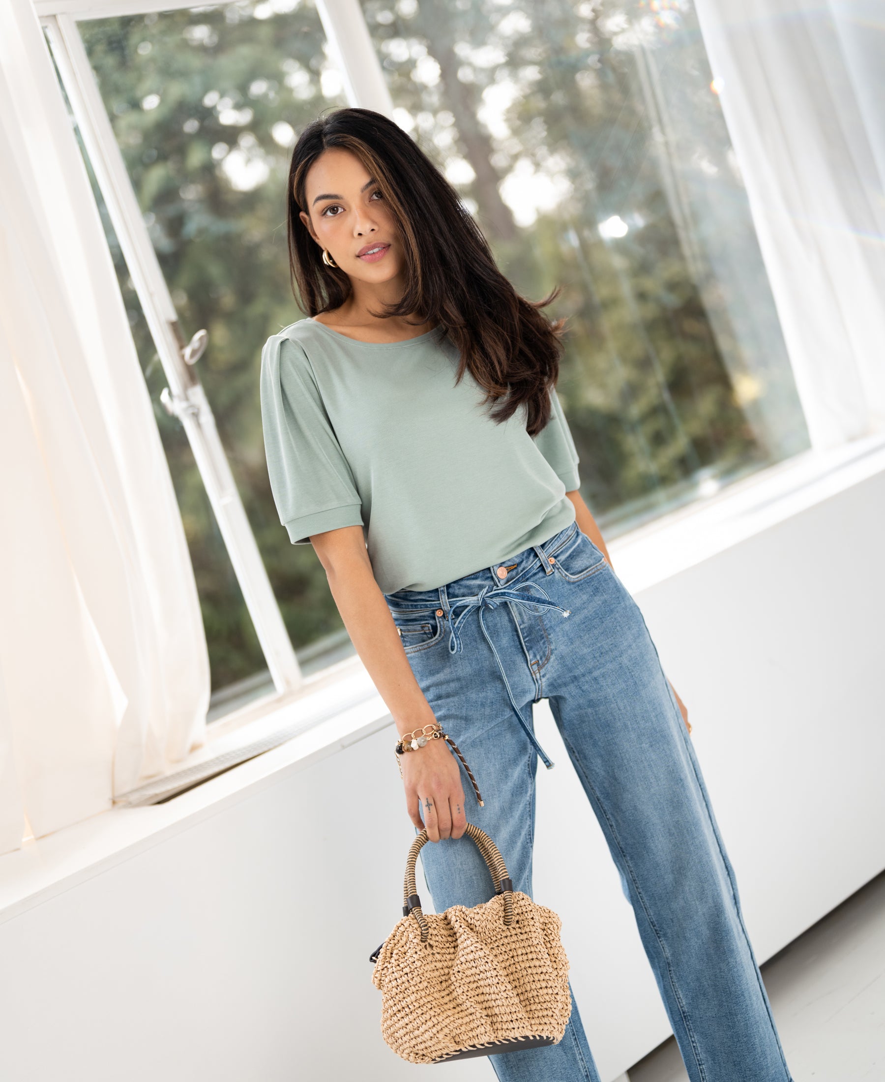 Woman wearing a light green ribbed T-shirt stands by a sunny window.