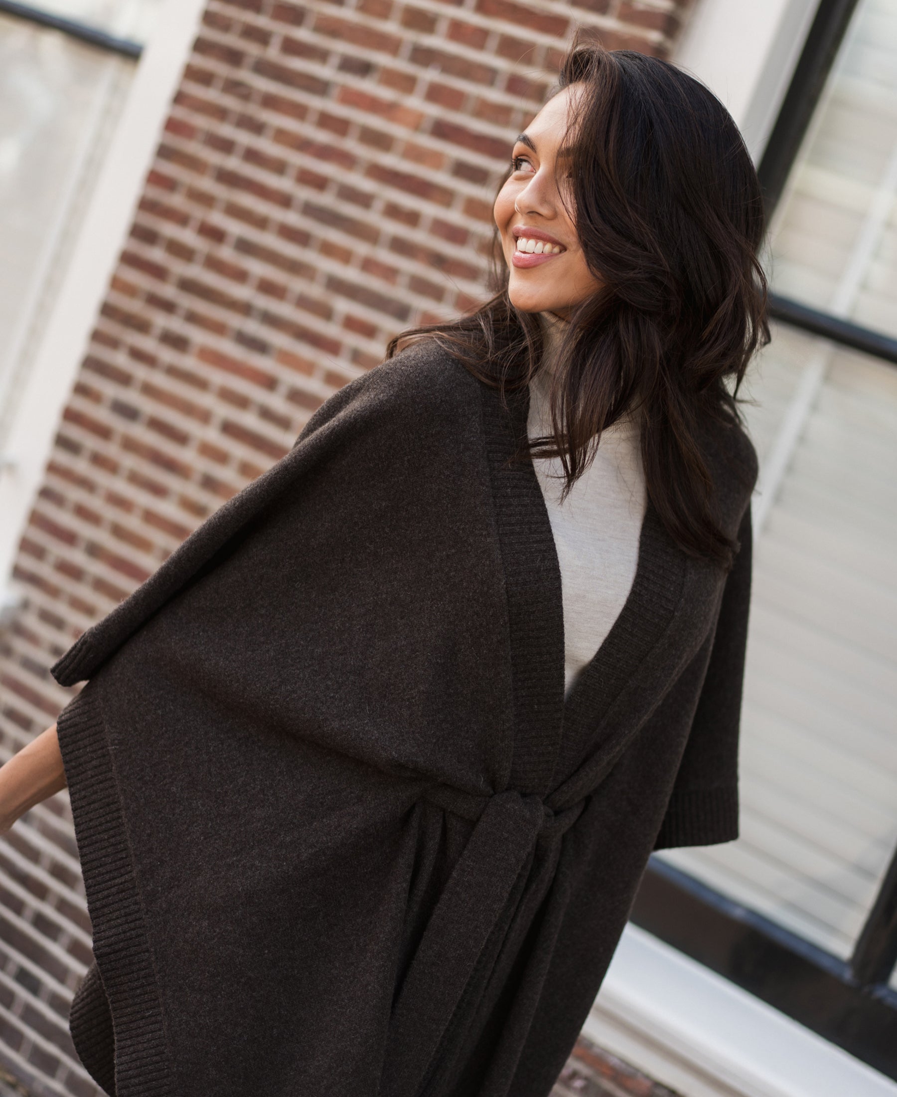 Woman wearing a pure chocolate brown merino wool poncho by PLEIN PUBLIQUE stands in front of a brick building.