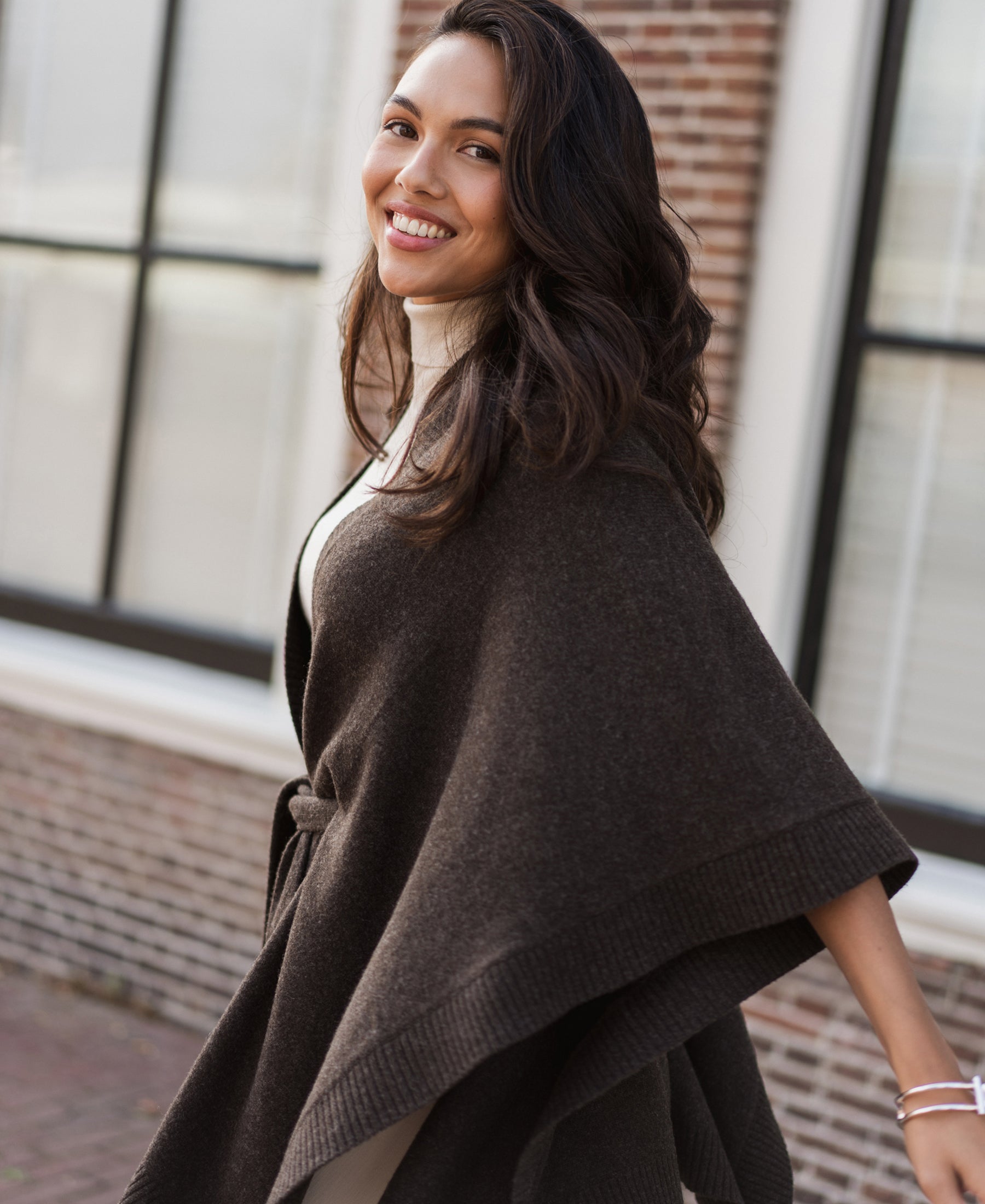 Woman wearing a chocolate brown merino wool poncho, standing by a brick building.