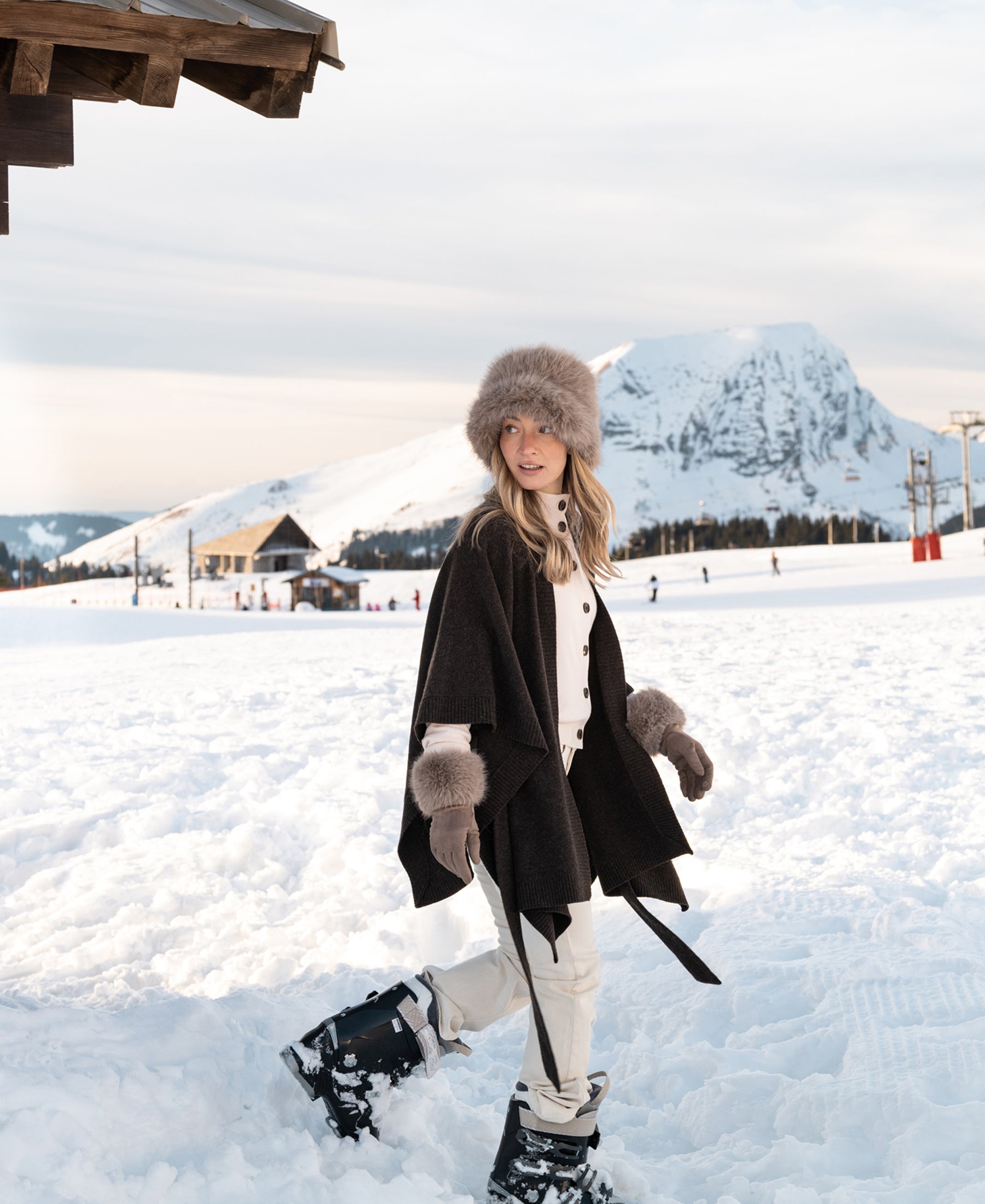 Woman wearing a pure chocolate brown merino wool poncho walks on snow with mountains and cozy cabins in the background.