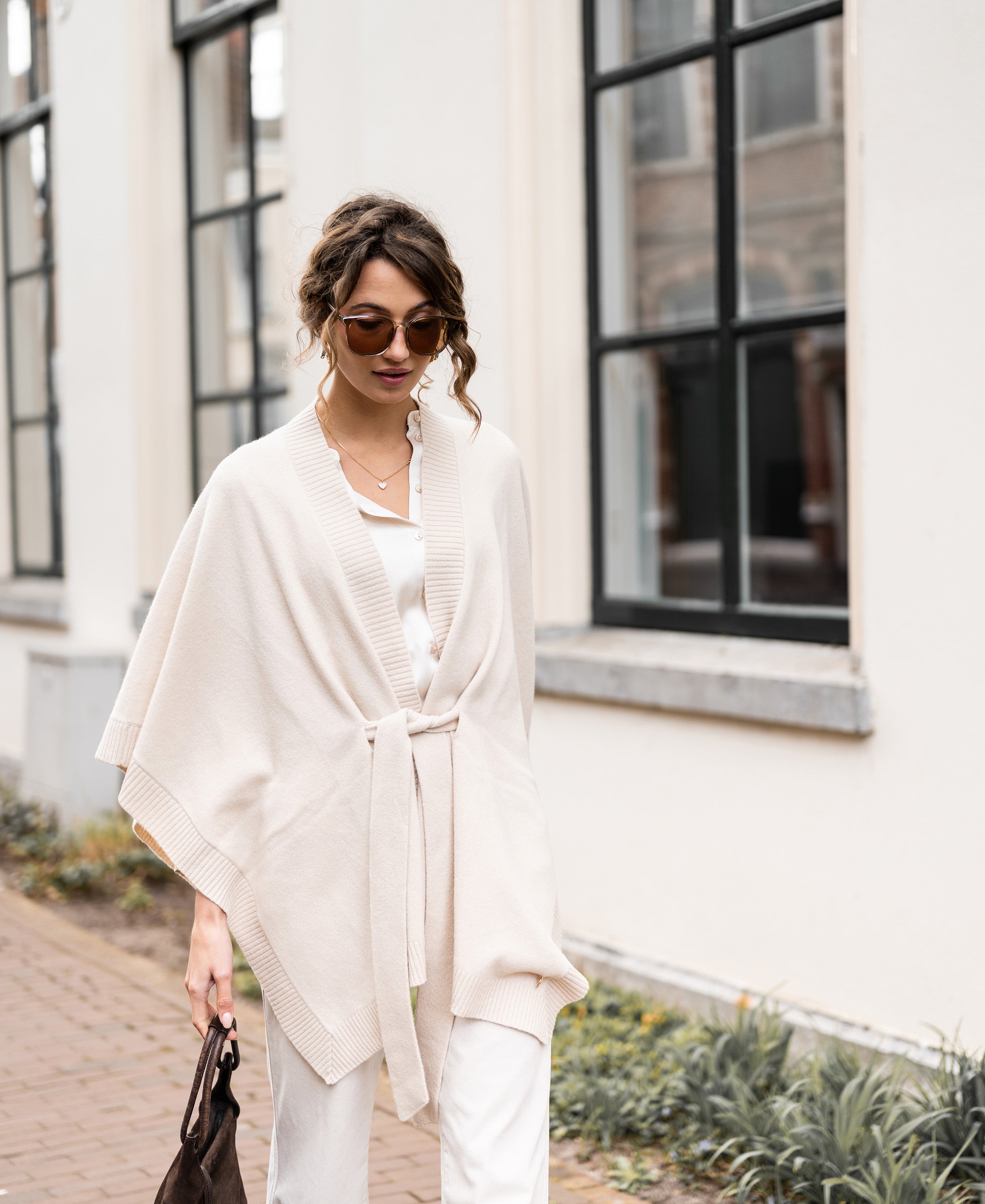 Woman wears light sand Merino wool poncho by a building with large windows.