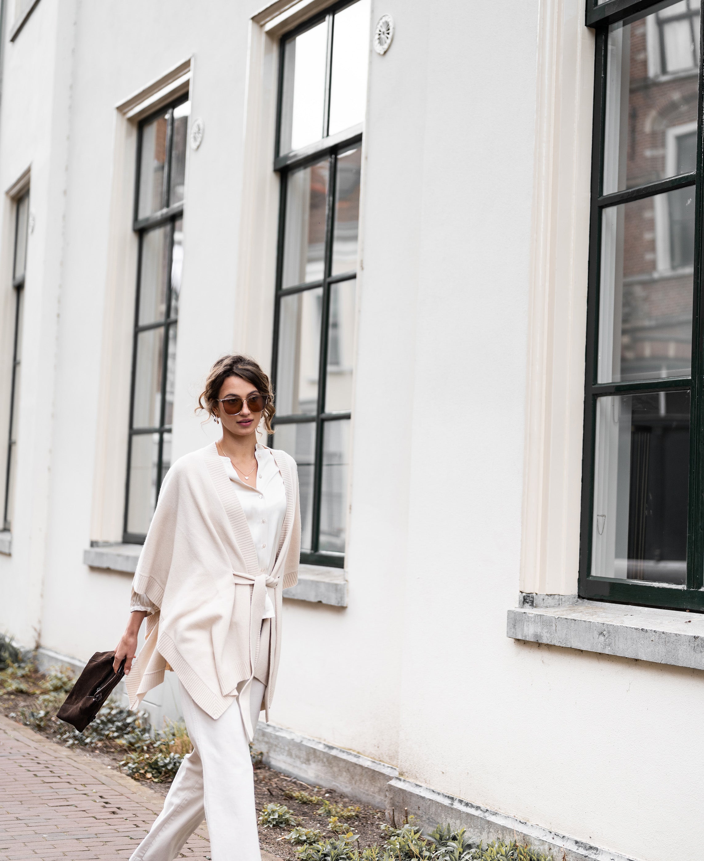 Light sand merino wool poncho with belt from PLEIN PUBLIQUE, shown on a woman standing by a white building.