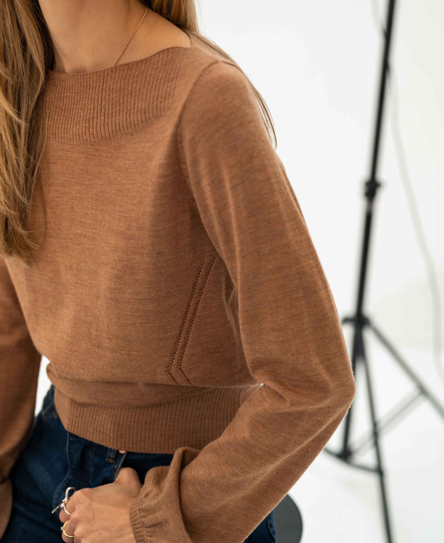 A woman wears a PLEIN PUBLIQUE boat neck sweater in chai tea color while sitting on a stool with studio lights behind her.