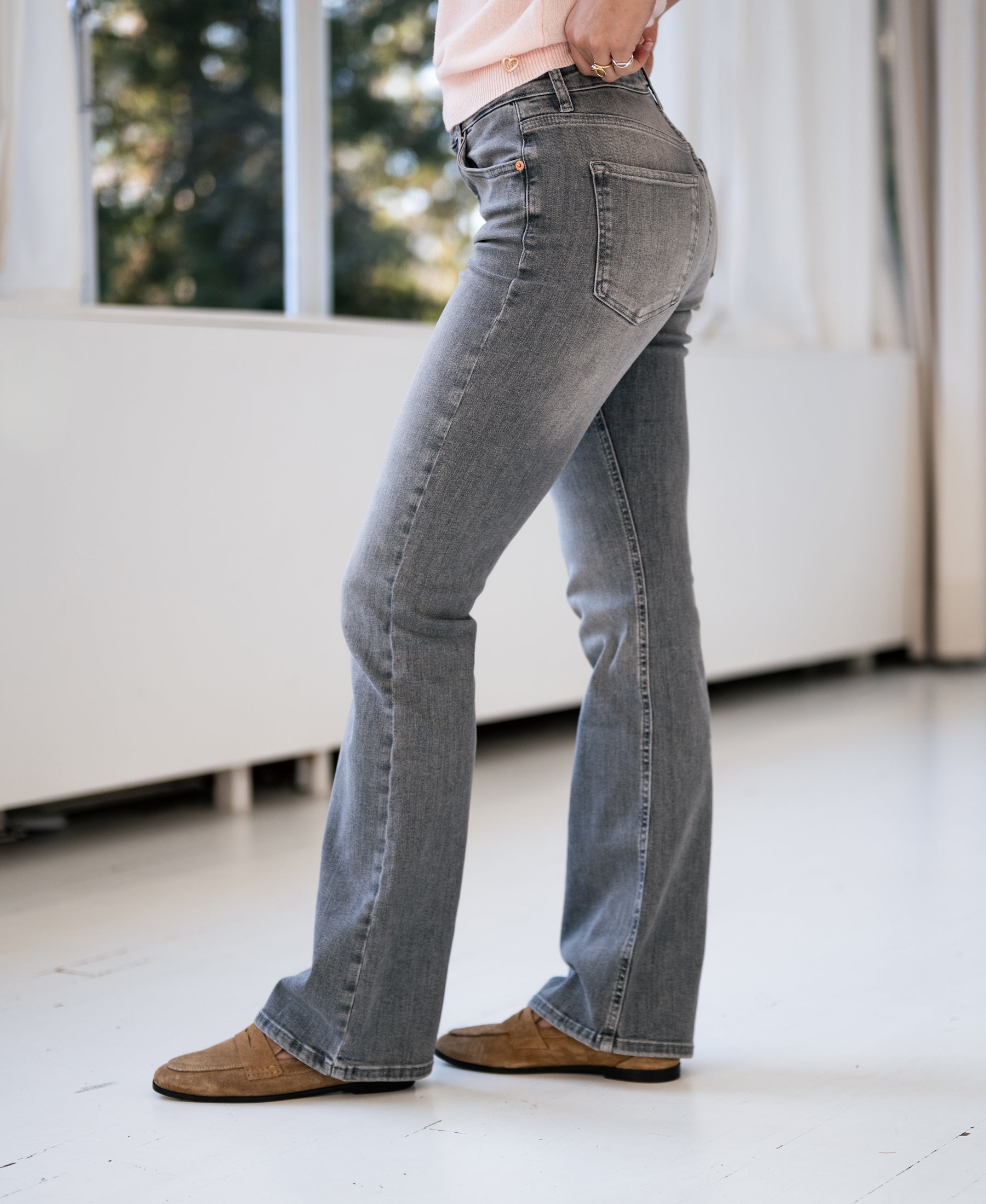 Person wearing grey stonewashed flared jeans stands indoors by a window with white curtains.