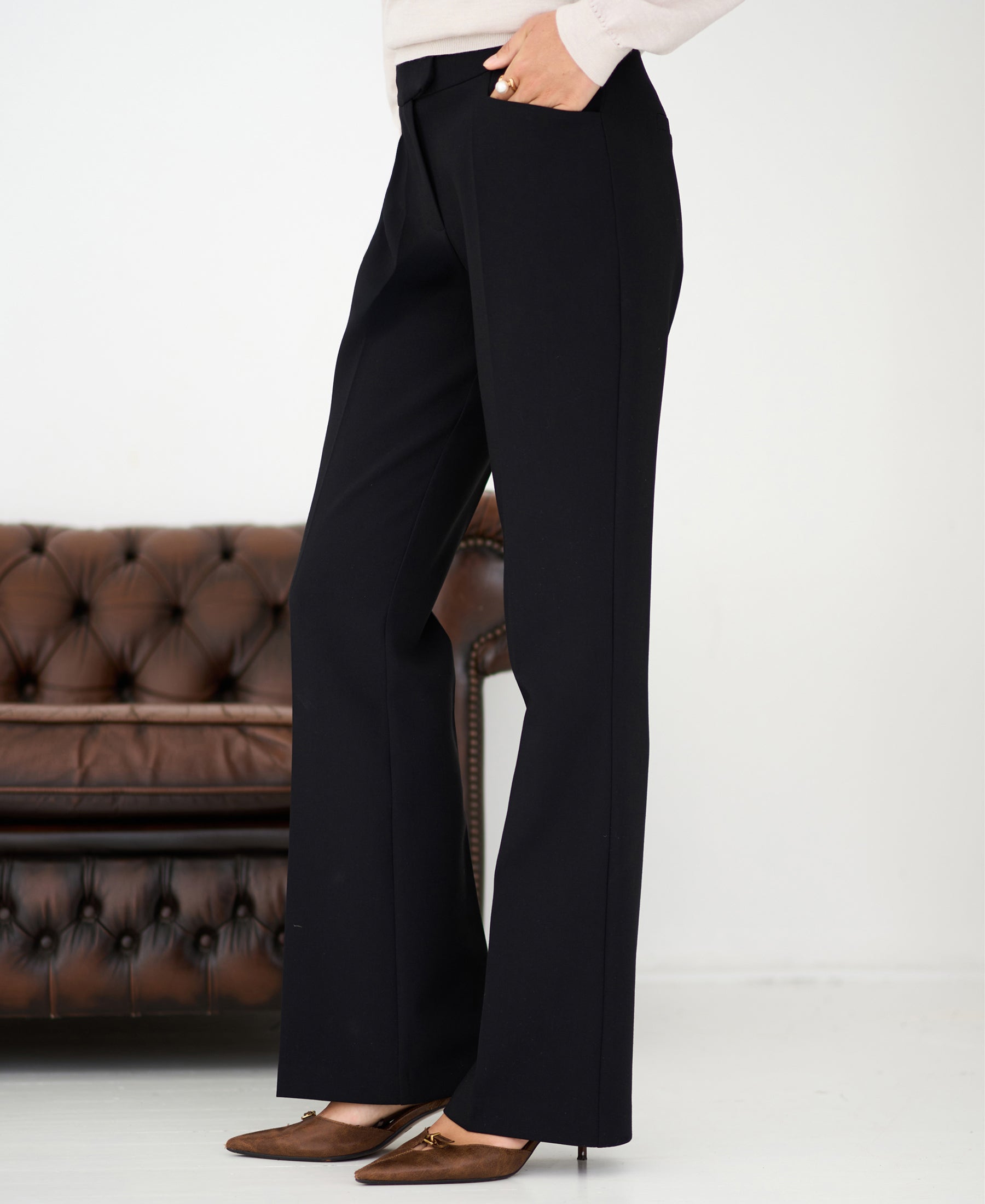 Model wears black flared pants by PLEIN PUBLIQUE, standing near a brown leather sofa.