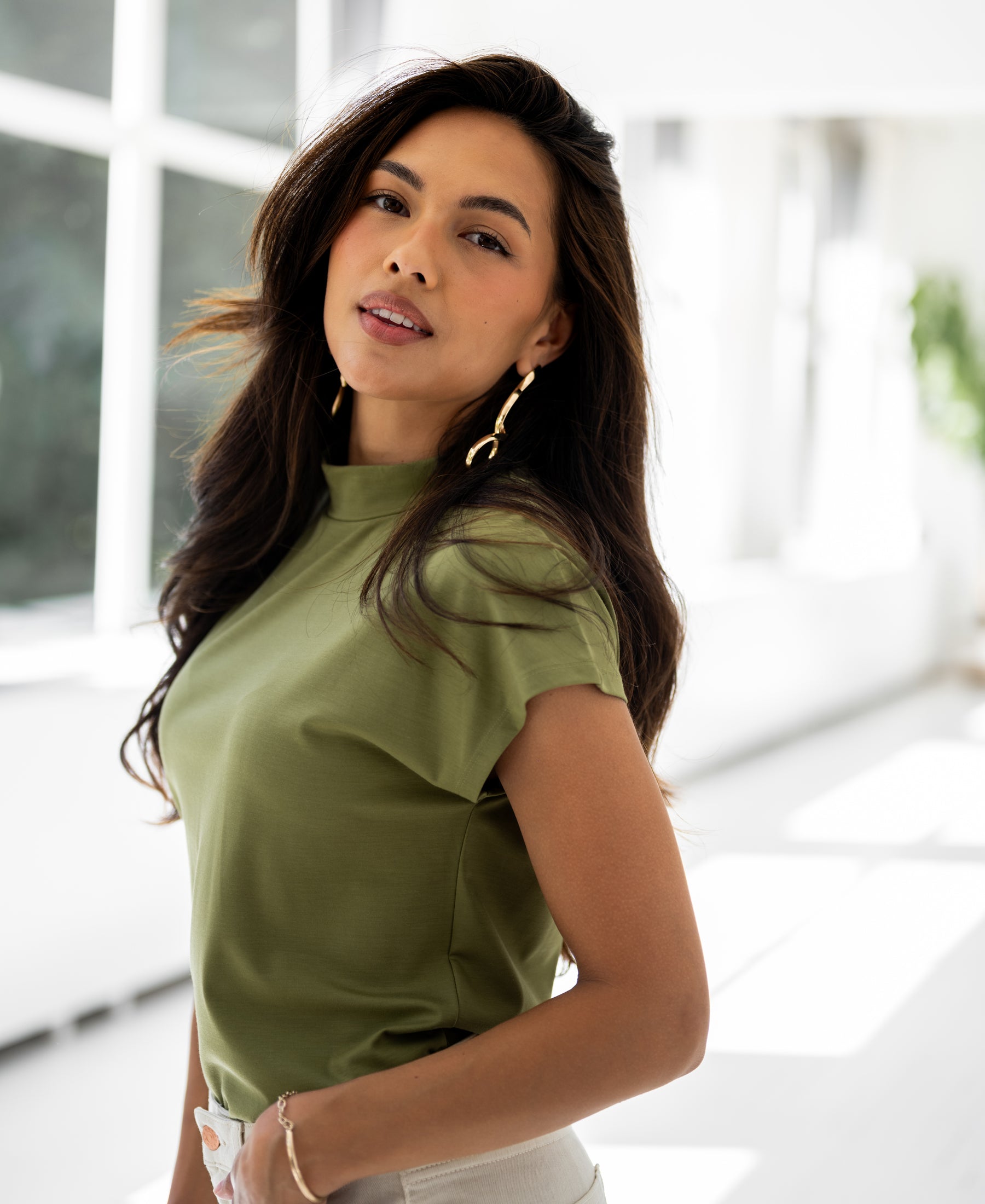 Olive green LA ROMY travel collared top by PLEIN PUBLIQUE shown in a bright, sunlit room.