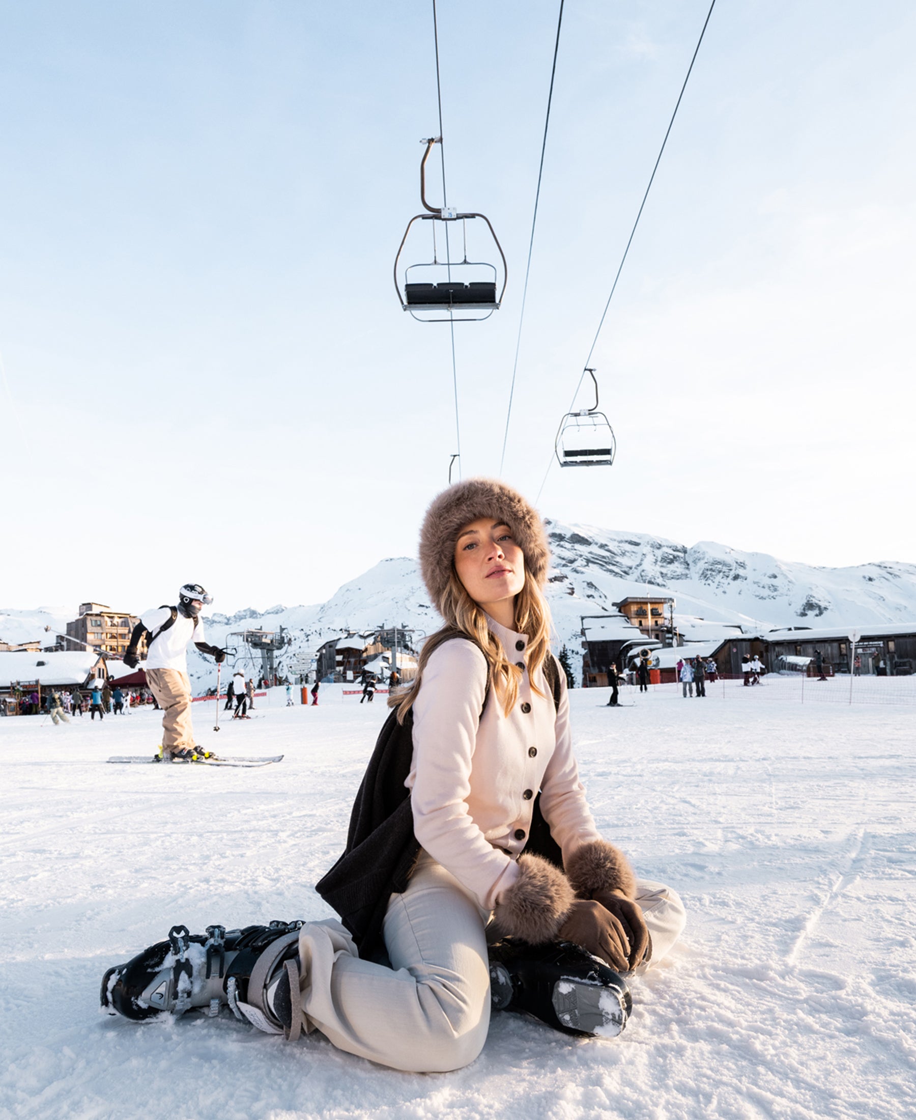 Woman wears blush-colored Merino wool turtleneck vest, kneeling in snow with ski resort mountains behind.