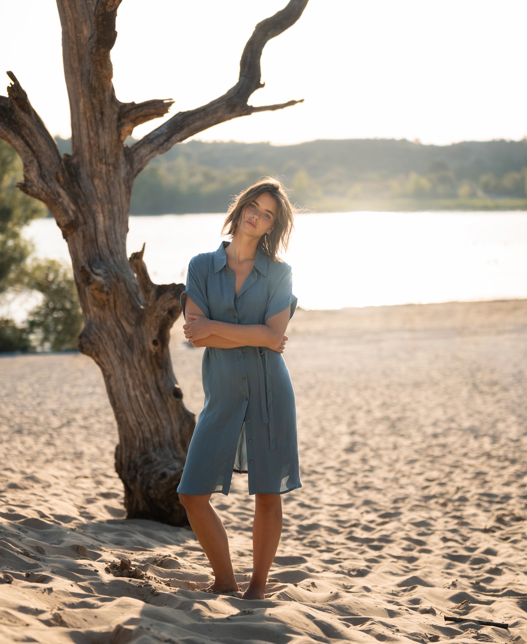 Cupro dress in Denim Cloud blue shown outdoors with tree, water, and hills in the background.