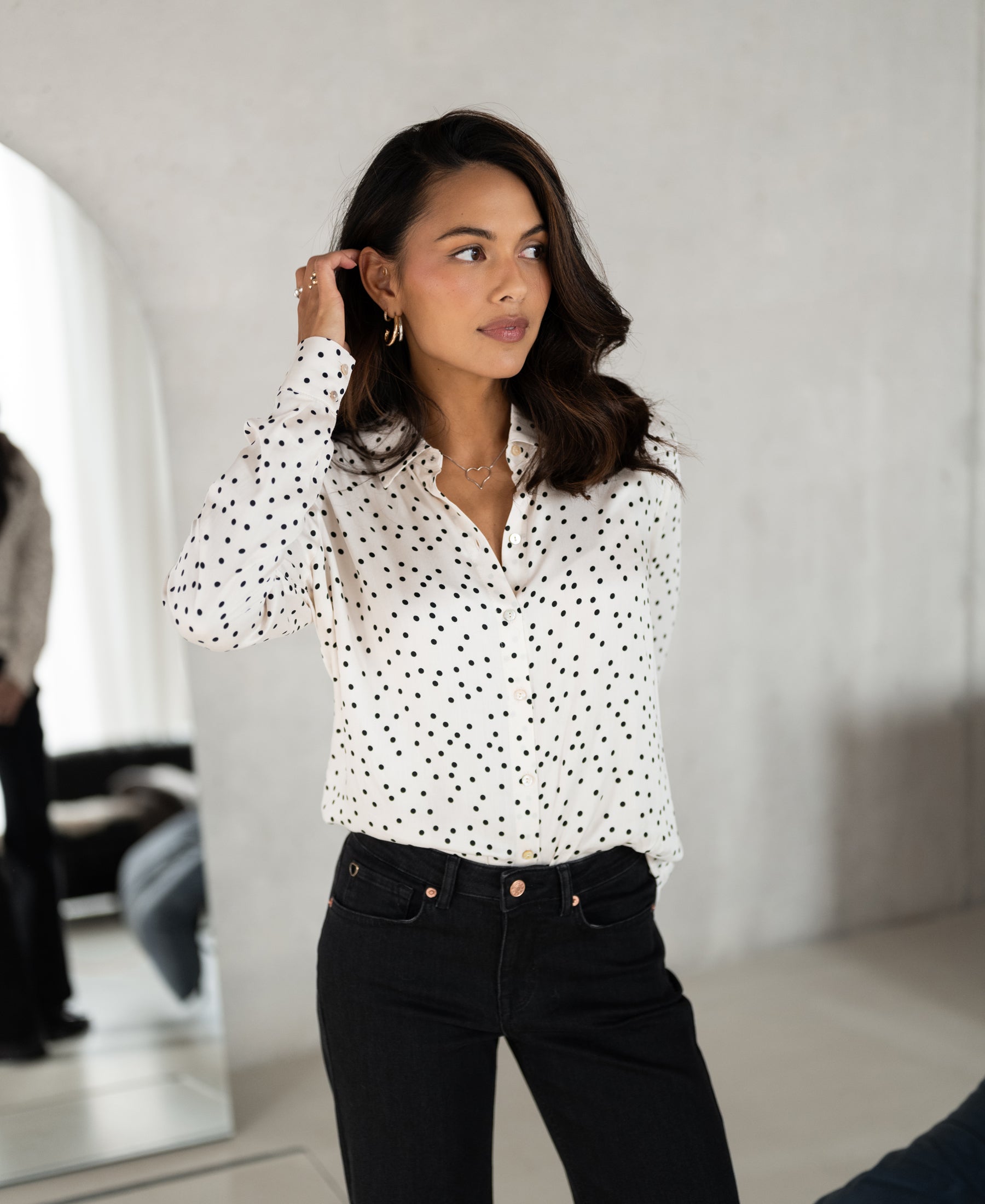 Woman wearing an ivory-black polka dot blouse by PLEIN PUBLIQUE LA SEZANNE stands near a mirror indoors.
