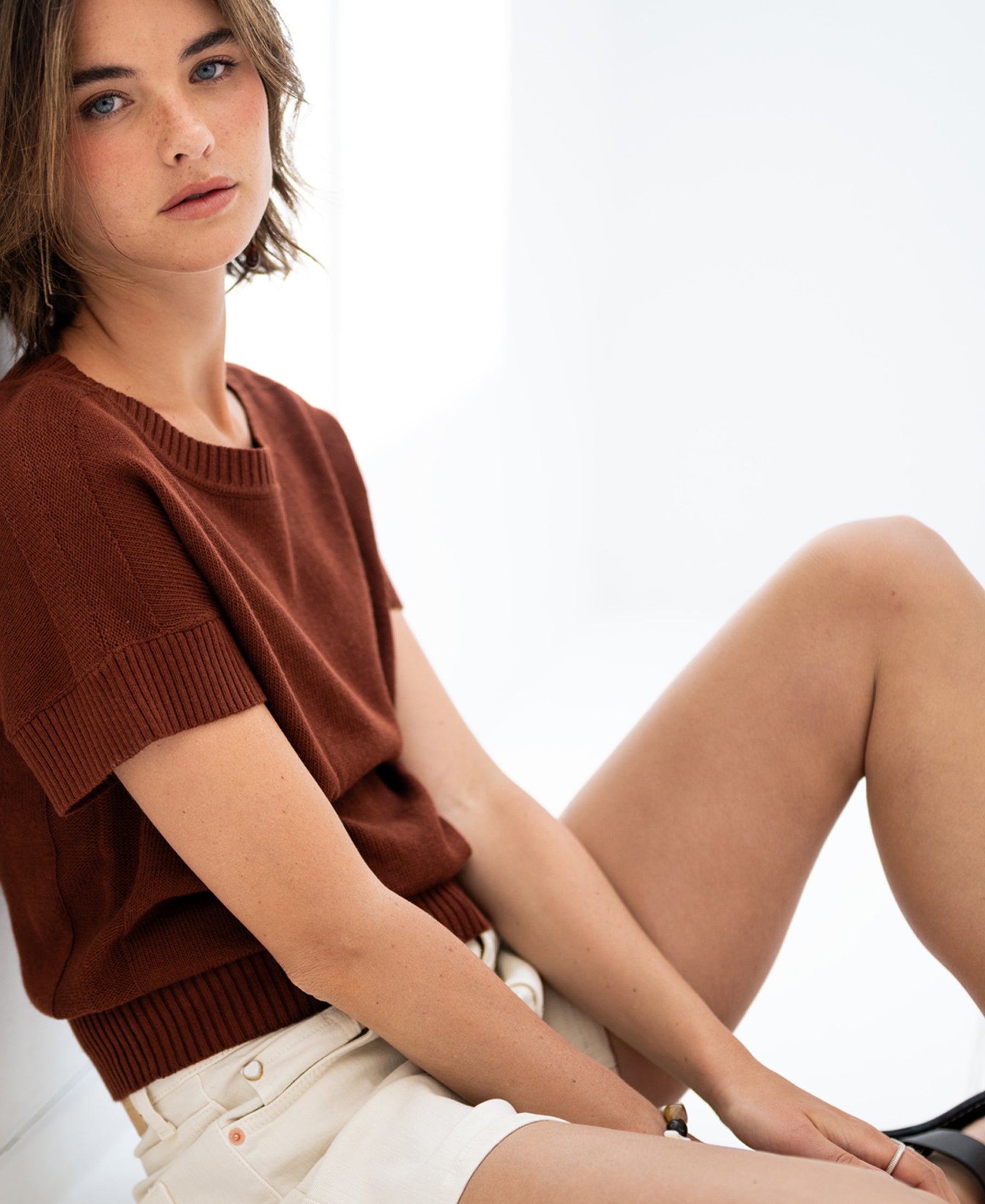 Woman wearing a linen summer sweater LE SOLEIL Sierra by PLEIN PUBLIQUE sits in a bright space.