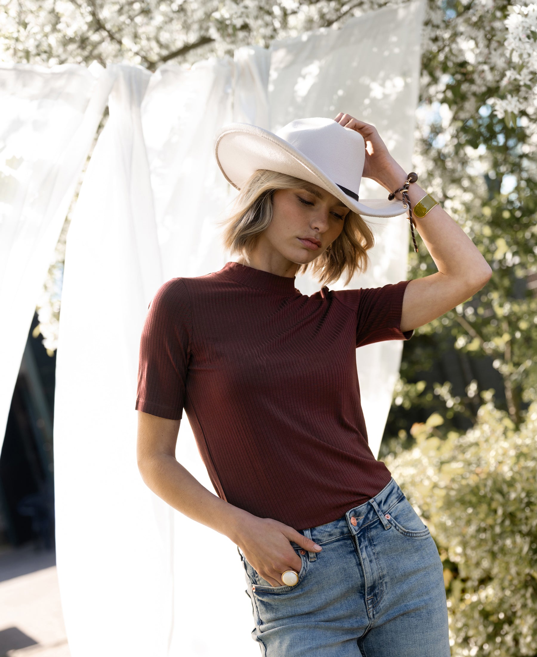 Woman wearing a chestnut bamboo turtleneck top stands outdoors by white drapes and blooming trees.