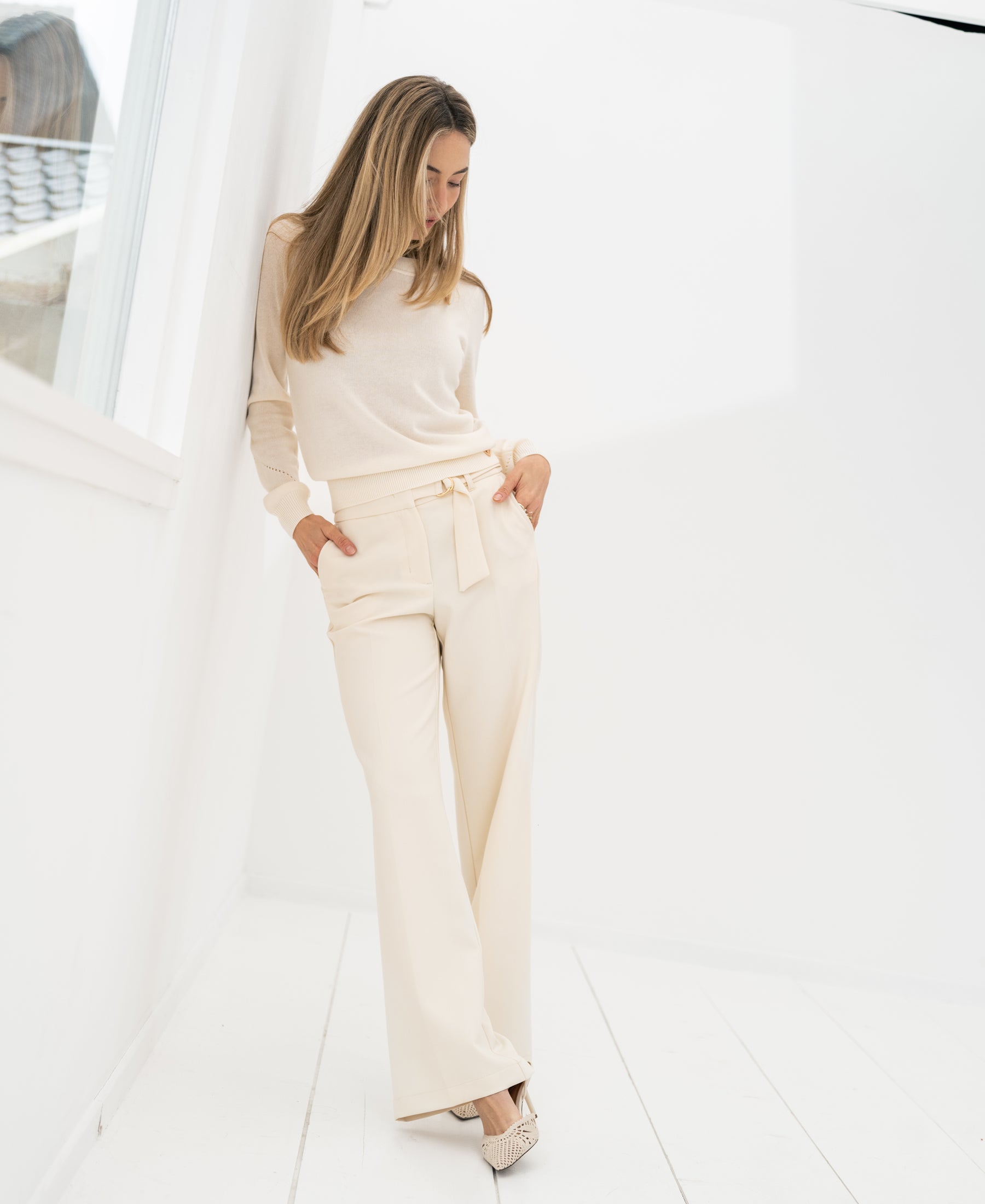 Woman wearing ivory Merino blend sweater leans against a white wall.
