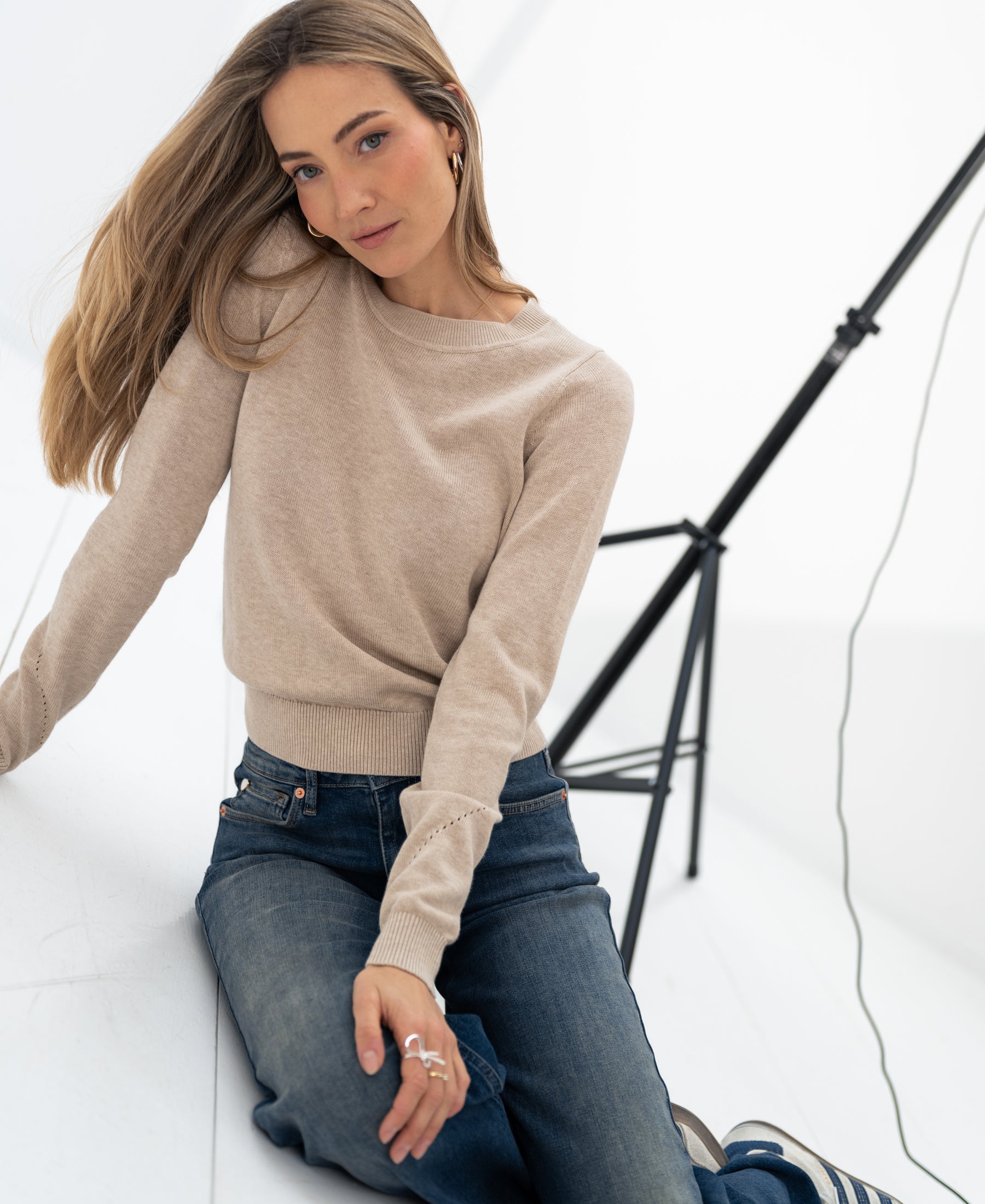Woman wearing a sand-colored merino wool sweater sits on a white floor under studio lights.