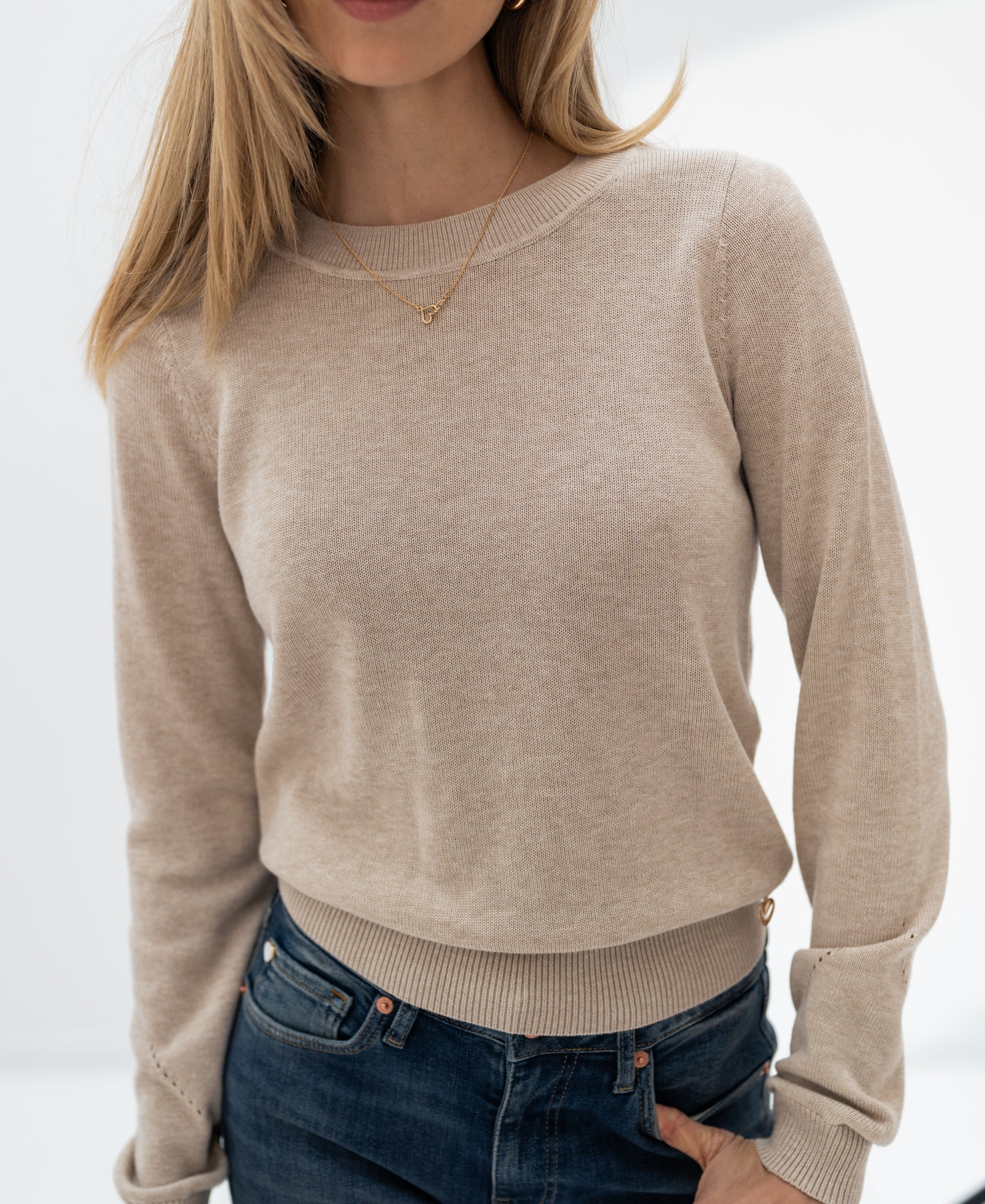 Merino wool sweater in sand color shown on a model standing with one hand in her pocket against a white background.