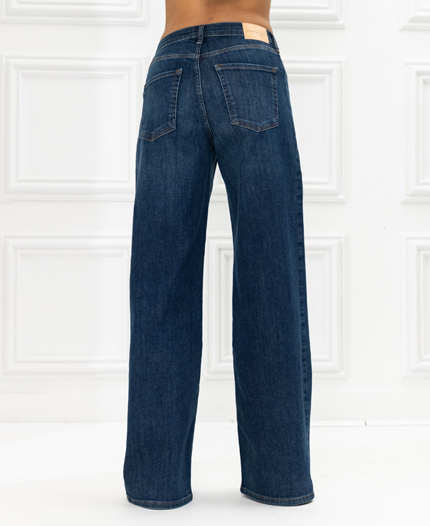 Wide leg blue jeans, shown from the back against a white paneled wall.