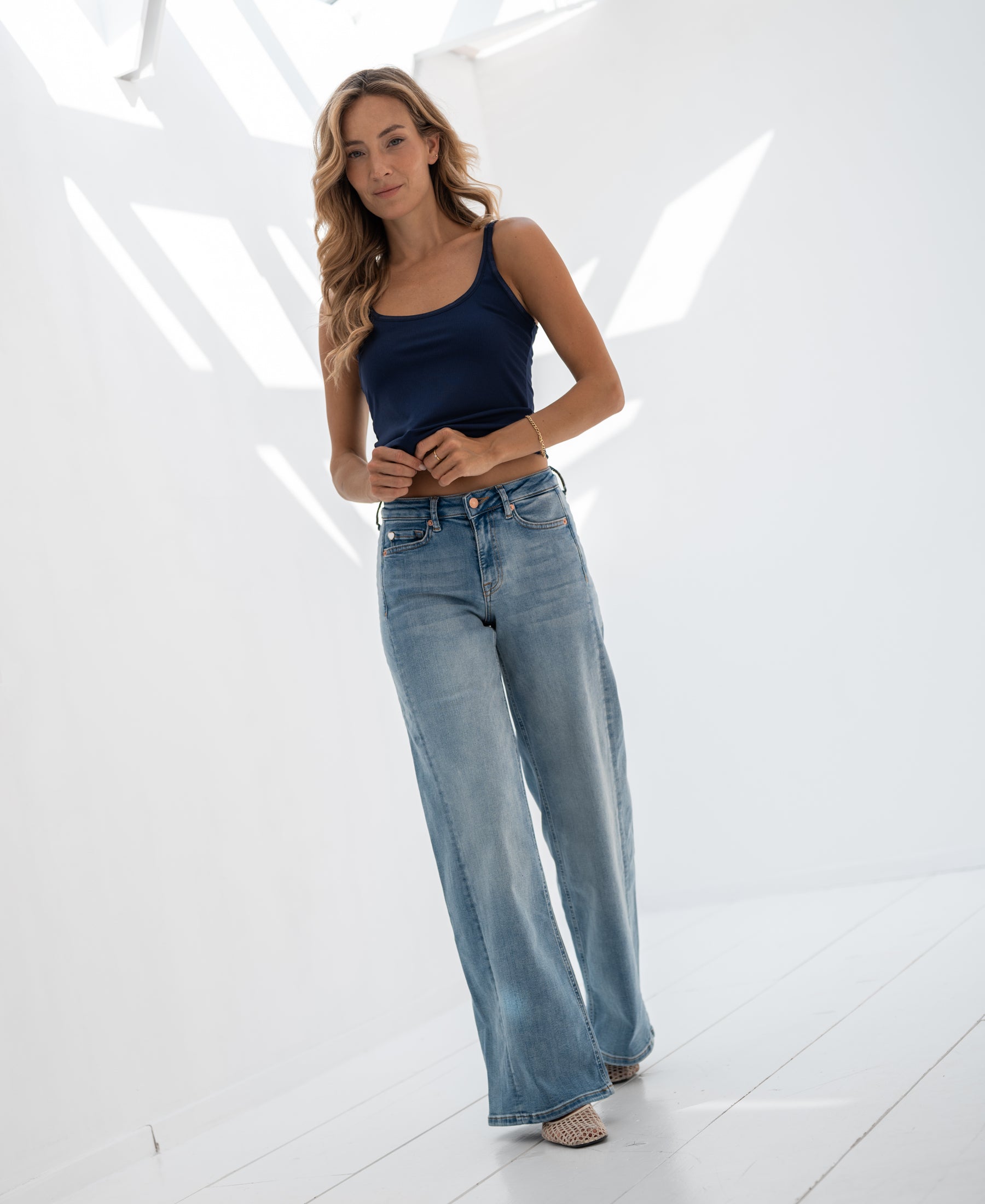 Woman wears navy silk cami top in a bright, sunlit white room.