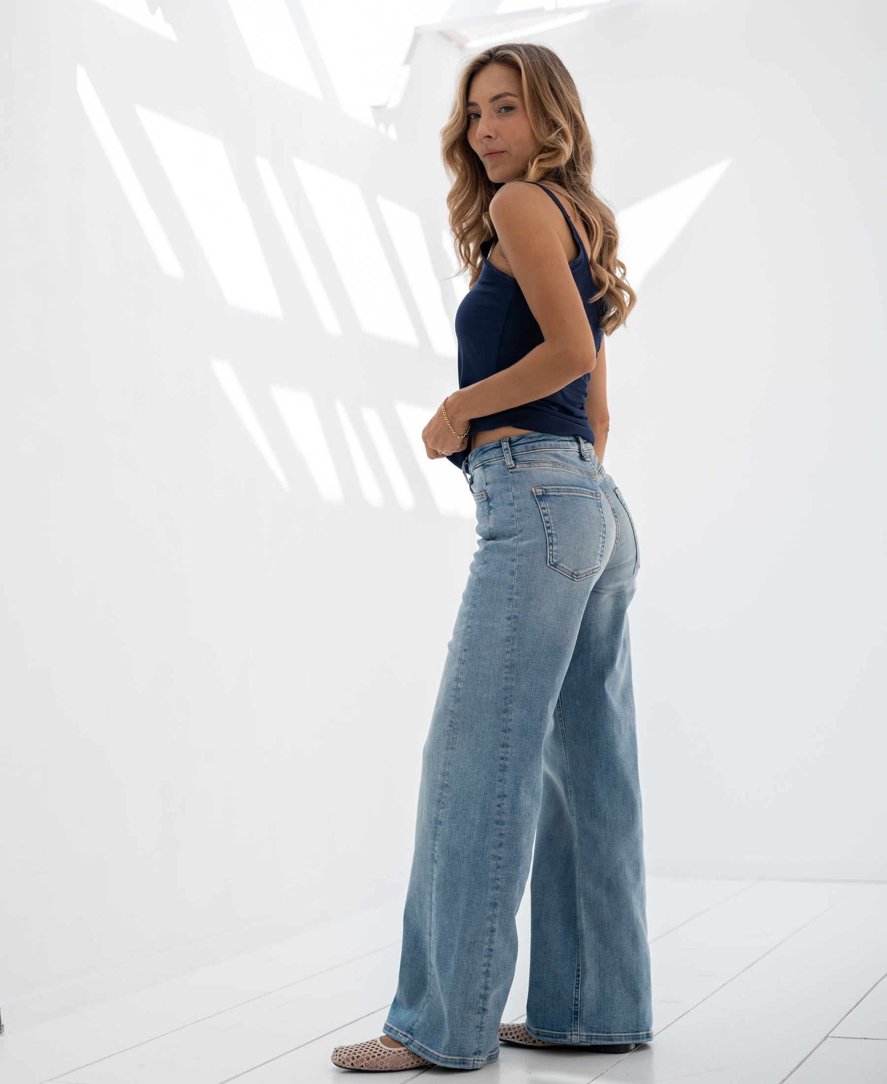 Wide leg jeans in light stone wash, shown on a model standing in a sunlit white room with shadows.