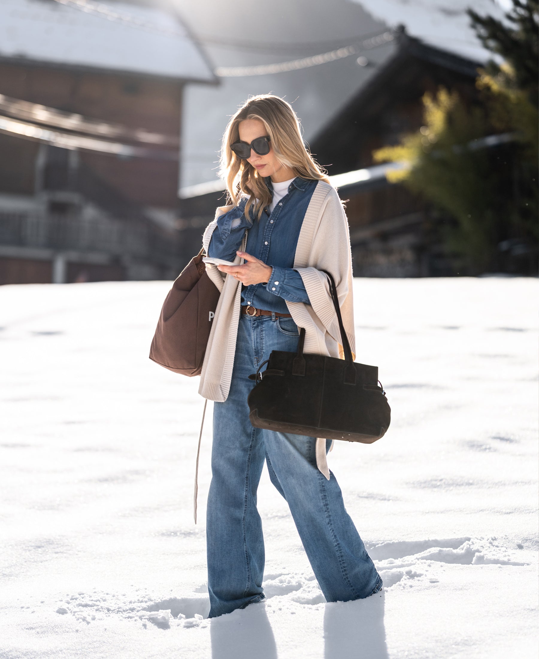 Woman wearing wide leg jeans in light stone color, walking outdoors on snow; cabins in the background.
