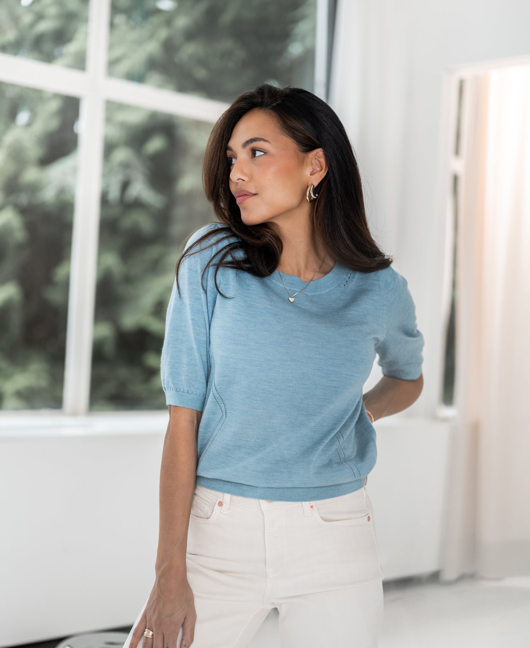 Woman wearing the PLEIN PUBLIQUE Sweater LA STEF in light blue, standing by a window and looking to the left.