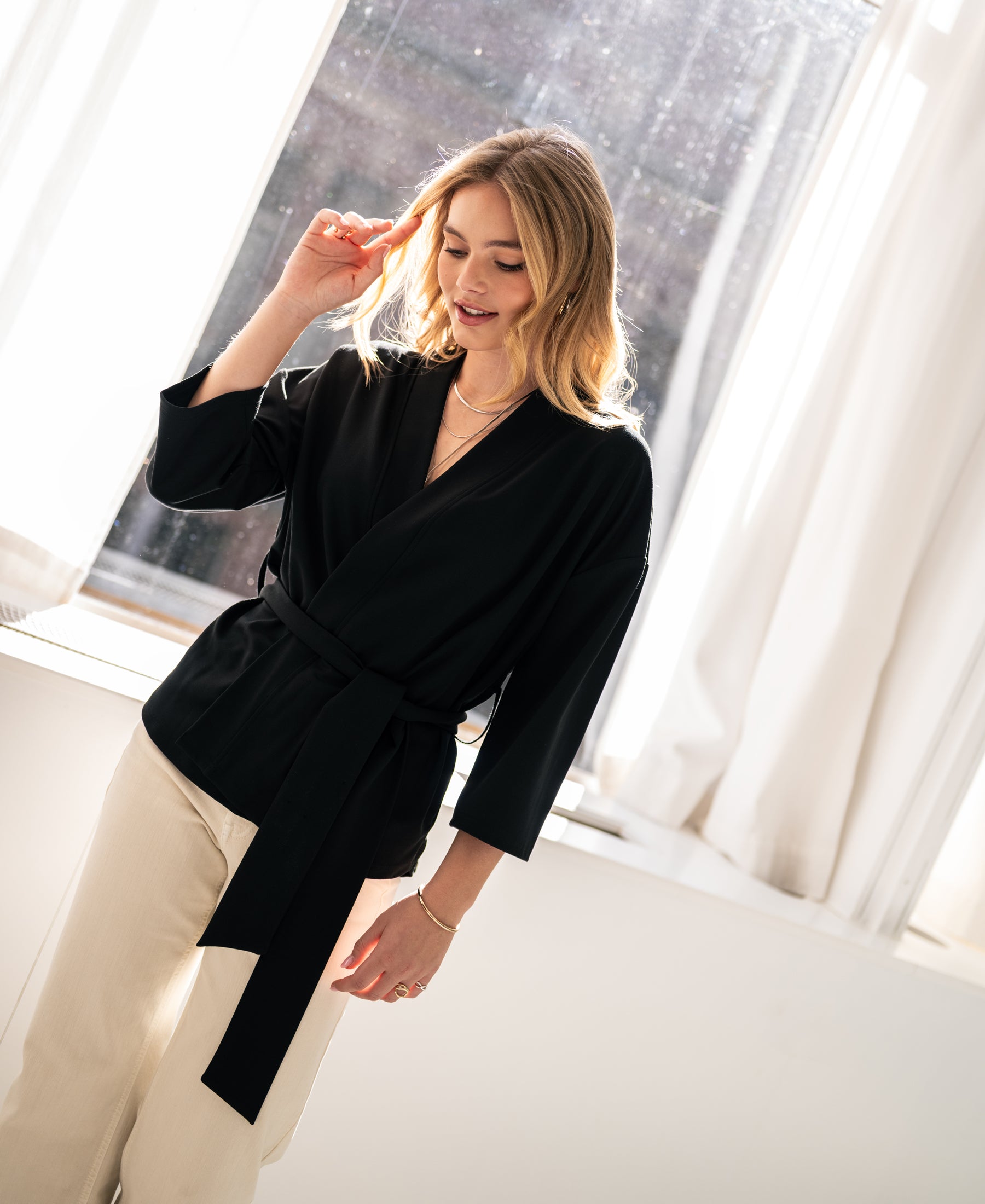 Woman wearing a black kimono wrap cardigan by PLEIN PUBLIQUE, standing by a sunny window.