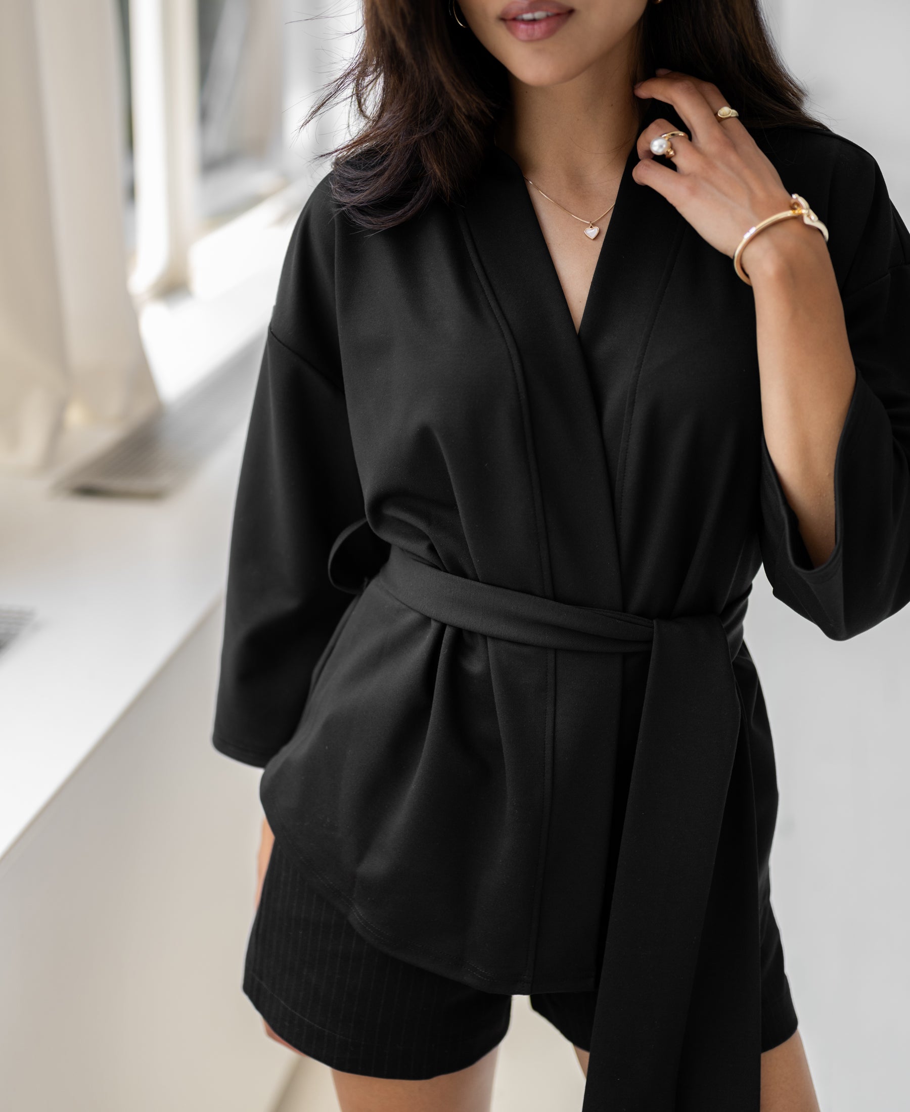 Woman wearing a black kimono wrap cardigan by PLEIN PUBLIQUE, standing by a bright window.