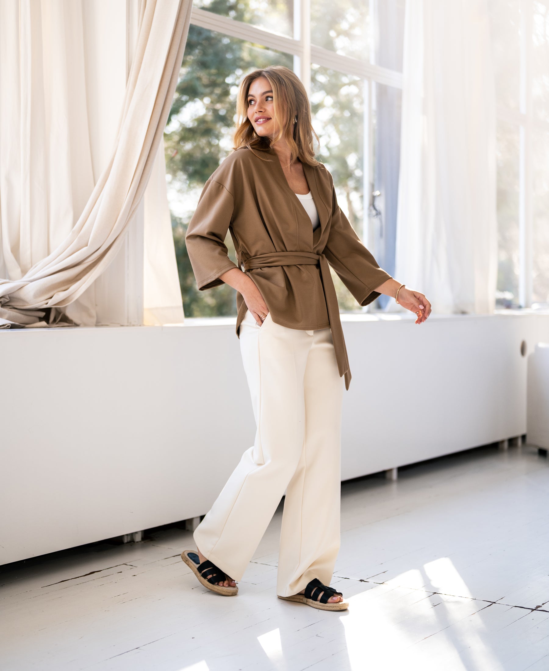 Woman wearing a PLEIN PUBLIQUE kimono wrap cardigan in dark sand, standing by a sunlit window.