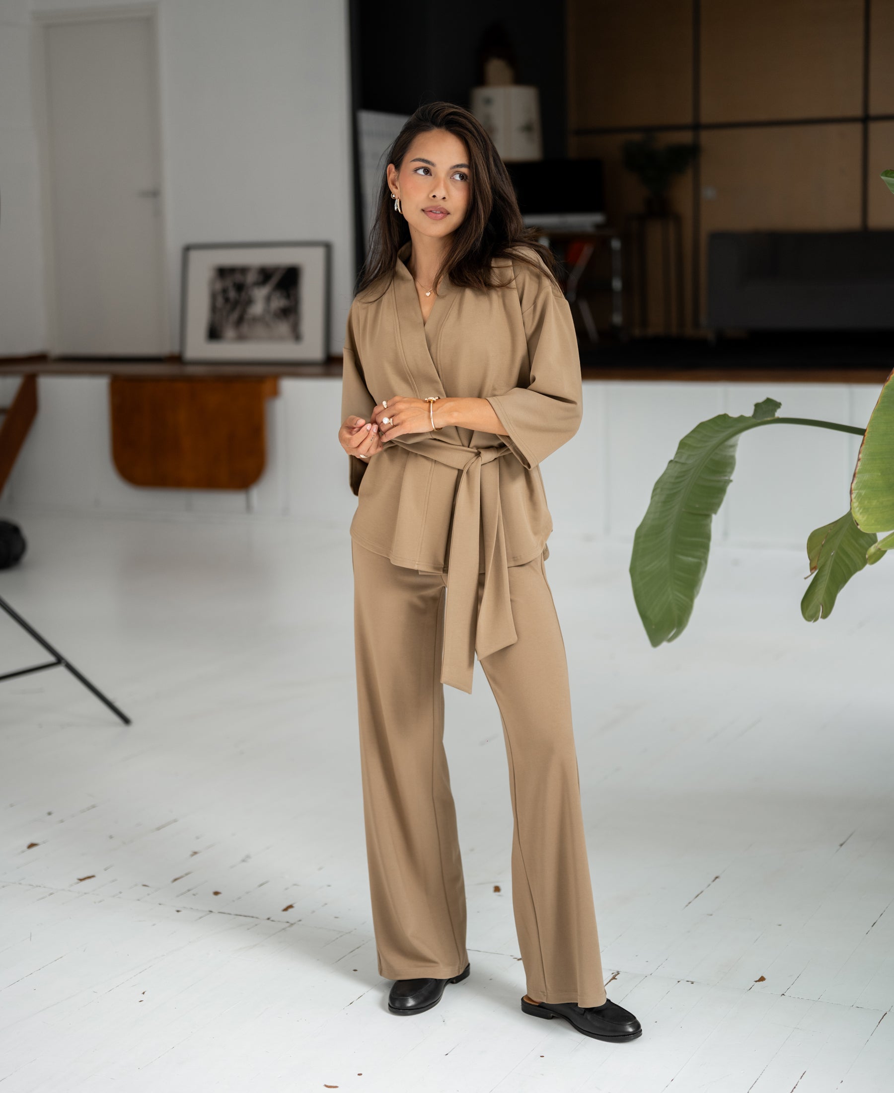 Woman wears dark sand-colored PLEIN PUBLIQUE travel pants, standing indoors.