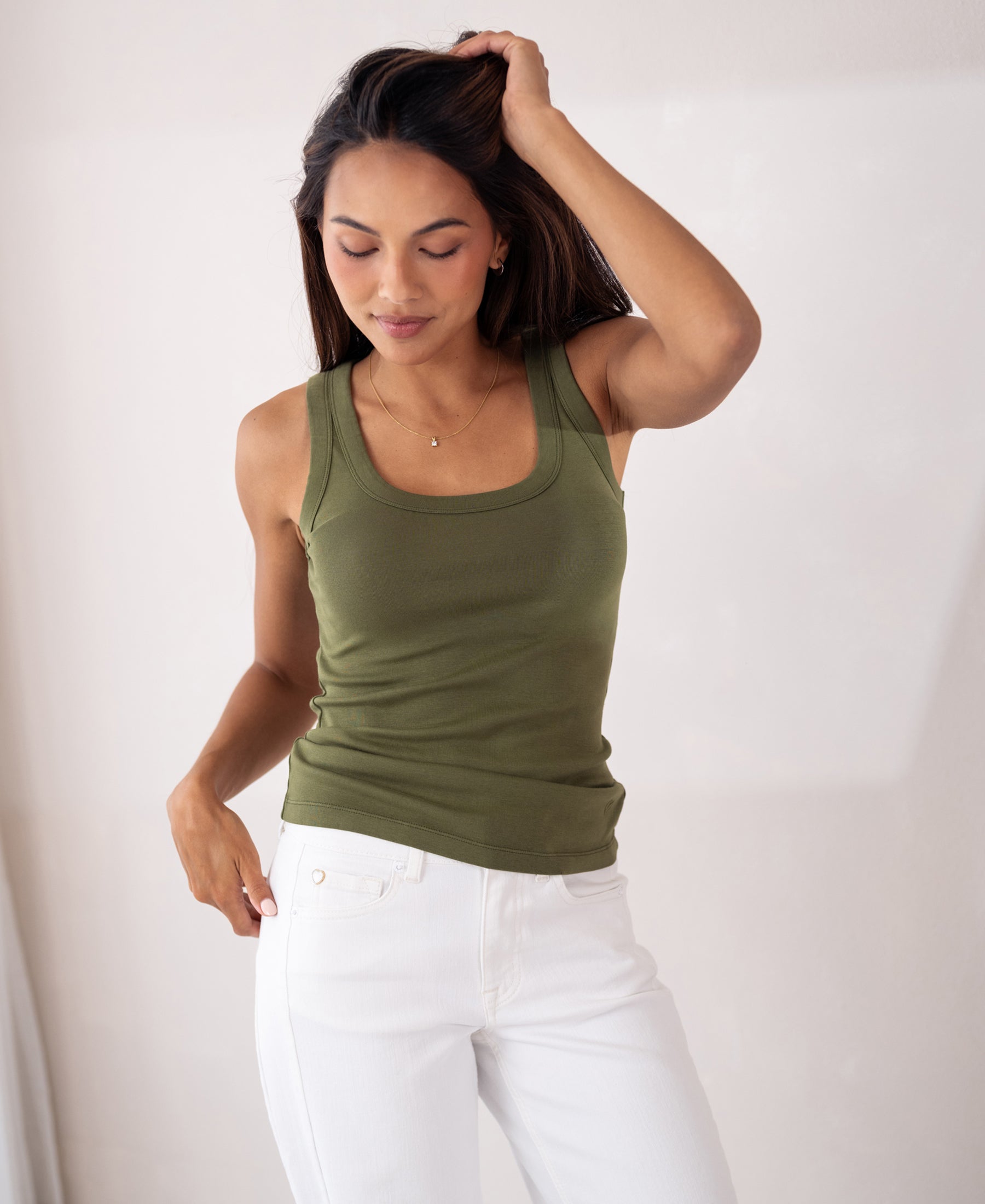 Woman wearing olive green LA TOULON Luxe & Soft top by PLEIN PUBLIQUE stands indoors, touching her hair and looking down.